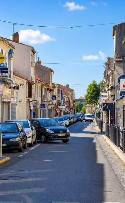Sunny commercial street in Mazargues, Marseille, lined with small shops and parked cars under a blue sky.