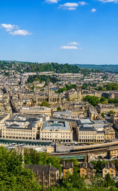 Aerial daytime view of Bath's honey-stone Georgian terraces and Bath Abbey in the valley, ringed by green hills.