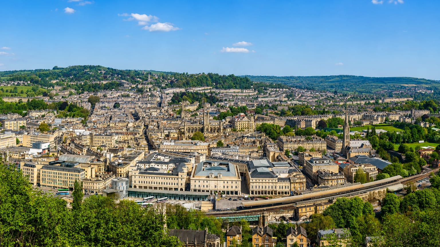 Aerial daytime view of Bath's honey-stone Georgian terraces and Bath Abbey in the valley, ringed by green hills.