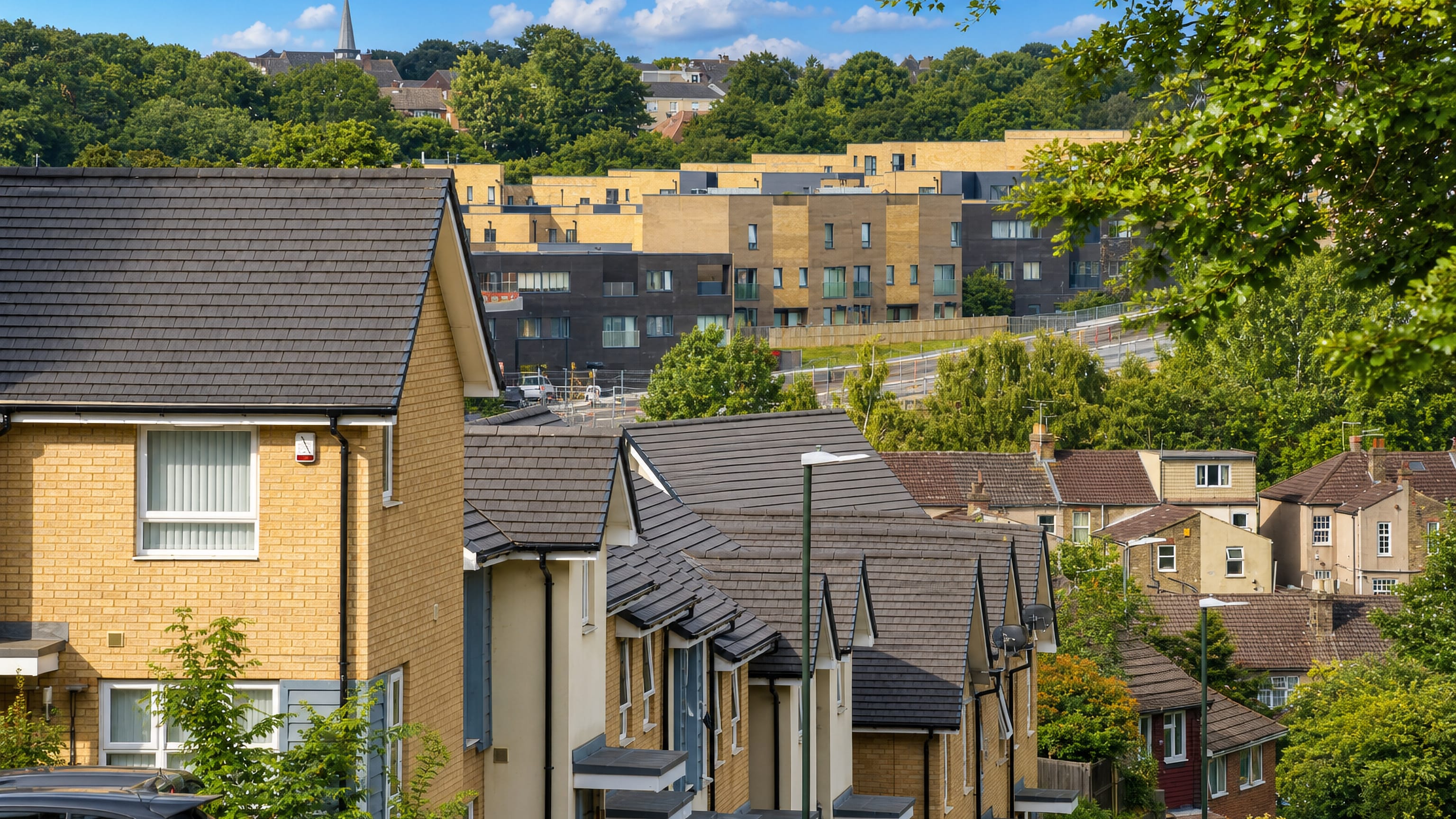Yellow-brick new-build terraces and older rooftops below a wooded ridge in Lessness Heath, Bexley.