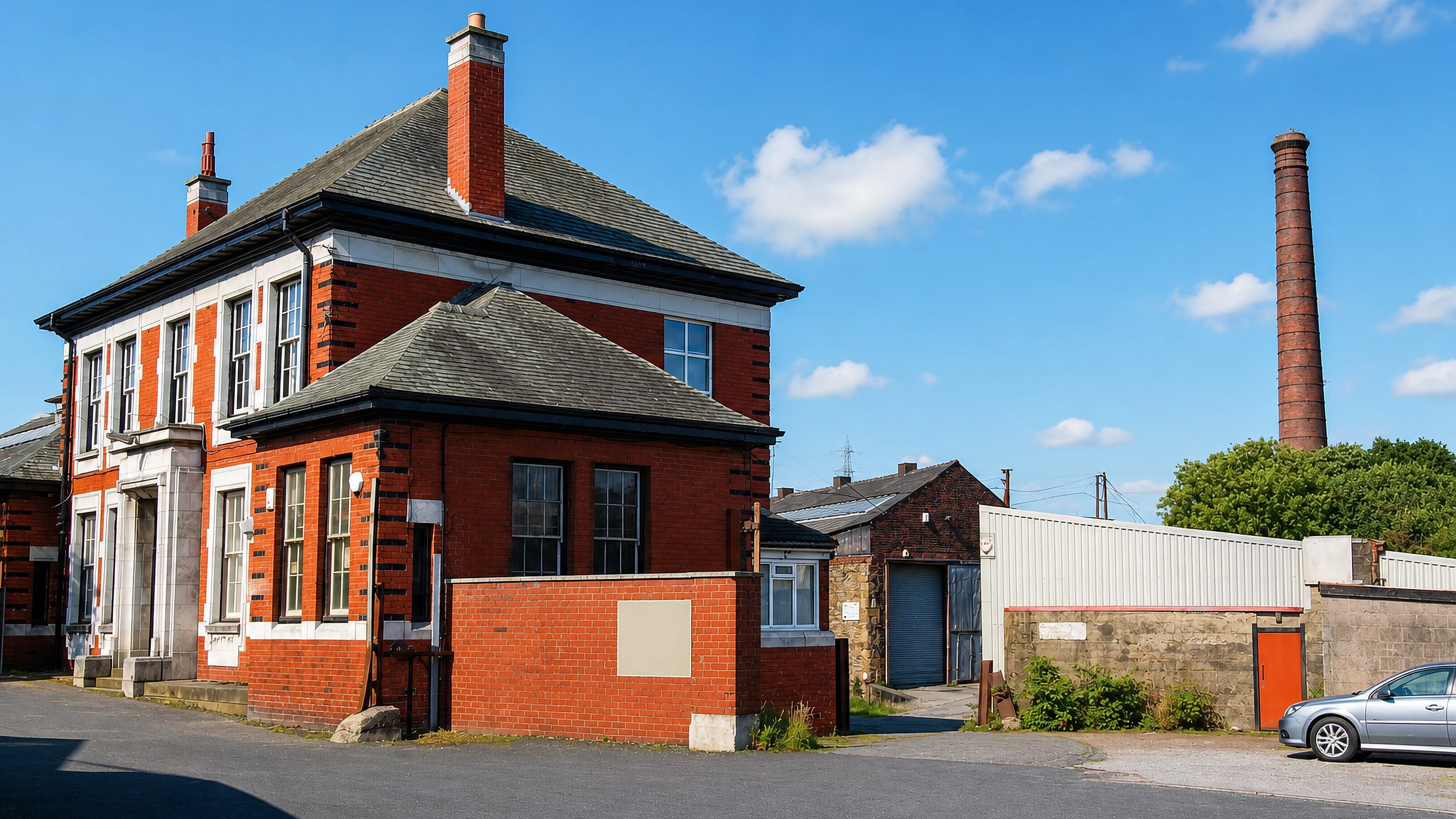 Red-brick Victorian building and a former mill chimney over an industrial yard in Breightmet, Bolton.