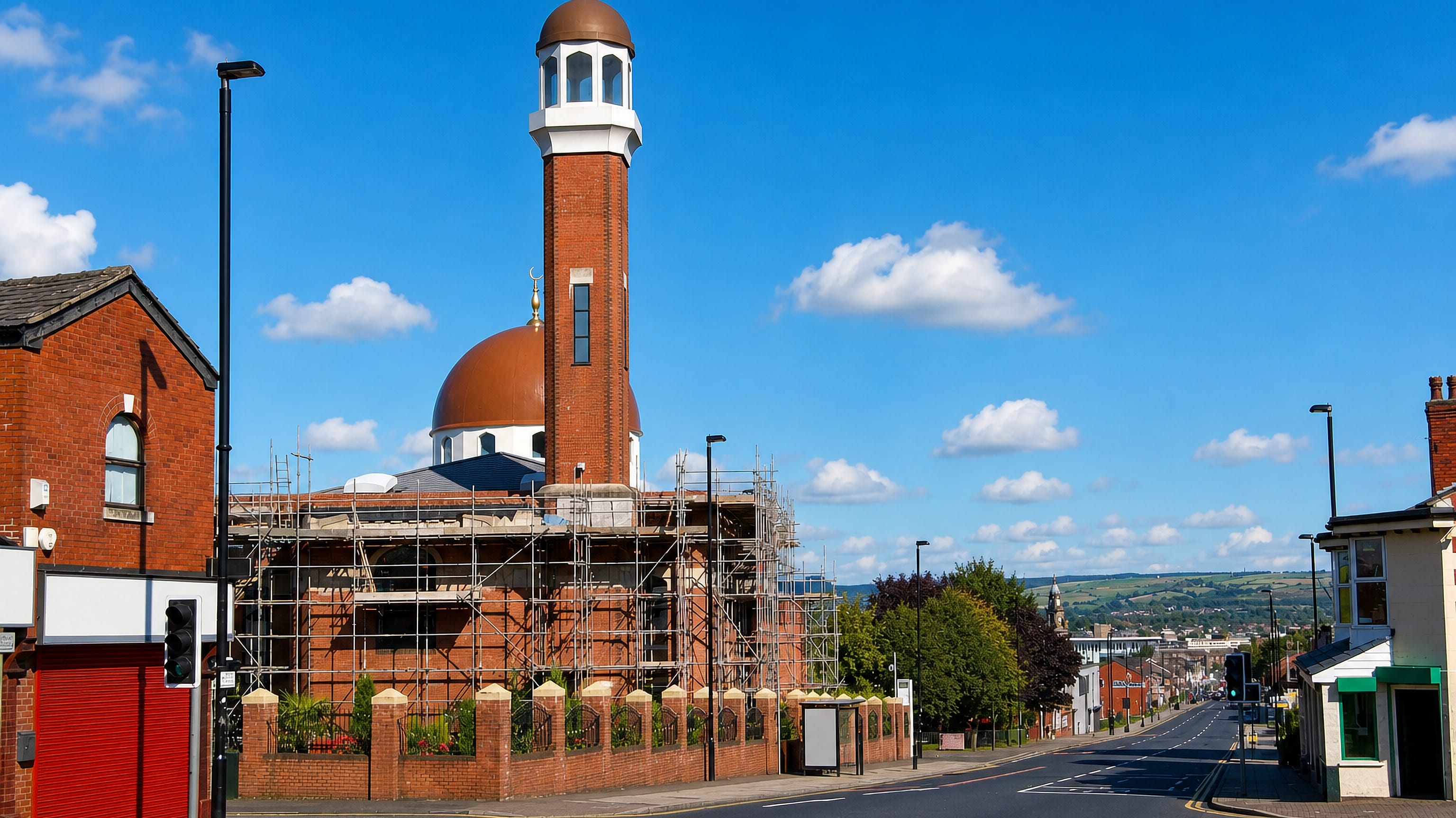 Brick mosque with copper dome and minaret on a main road through Daubhill, Bolton, with moorland hills beyond.