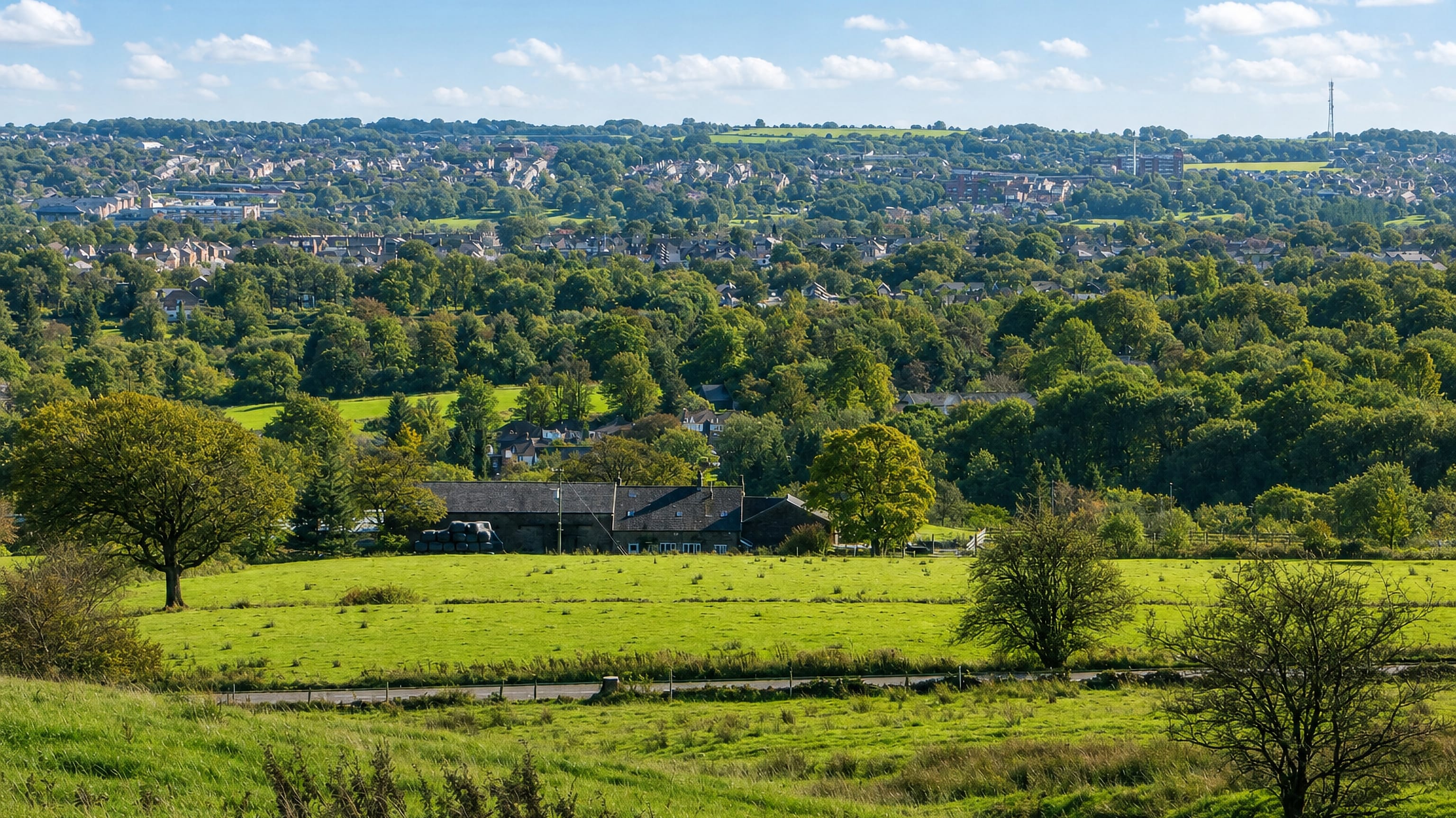 Green fields above Bolton's rooftops seen from the moor edge at Smithills, with a hilltop mast beyond.