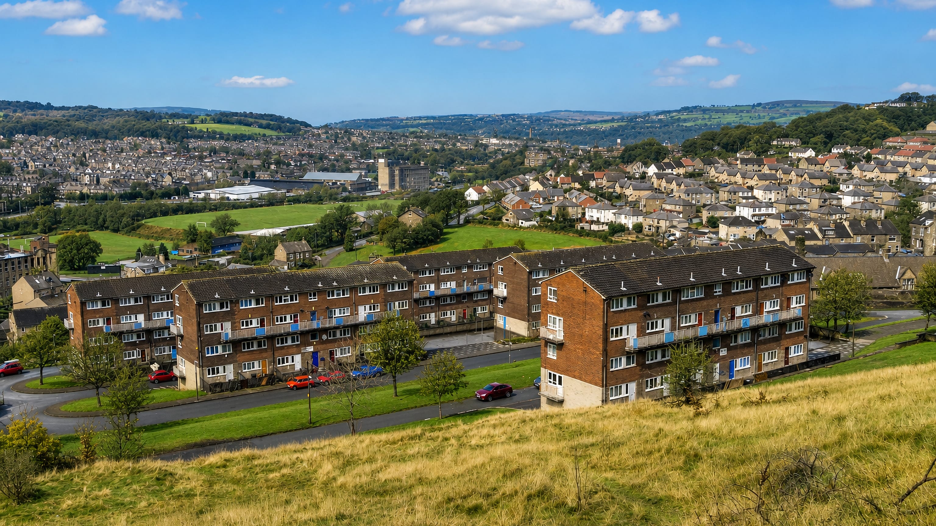 Brick maisonette blocks and stone terraces on the hillside at Bolton, Bradford, with Pennine hills beyond.