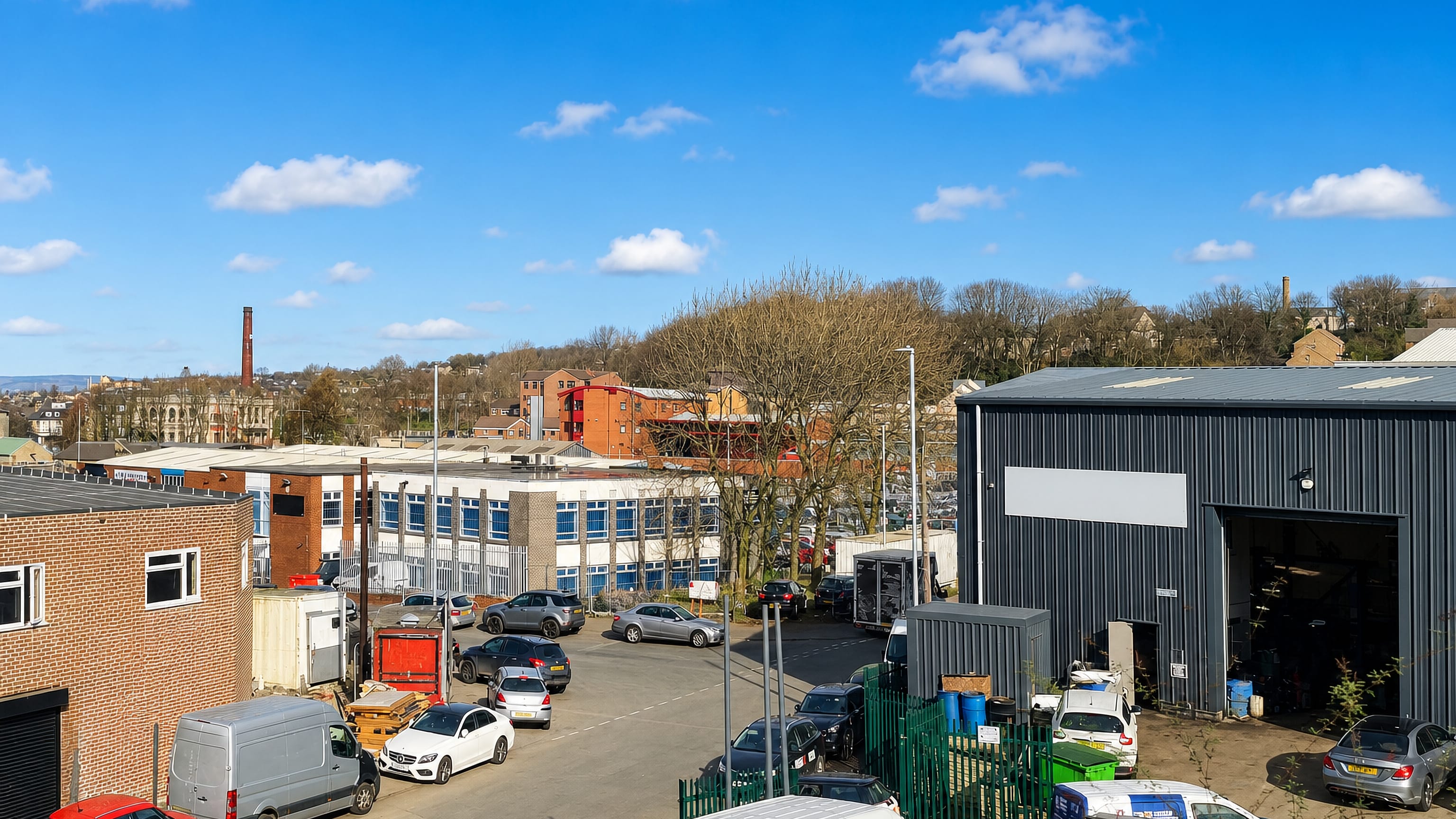 Industrial units, parked vans and stone terraces below a wooded ridge and mill chimney in Bowling, Bradford.
