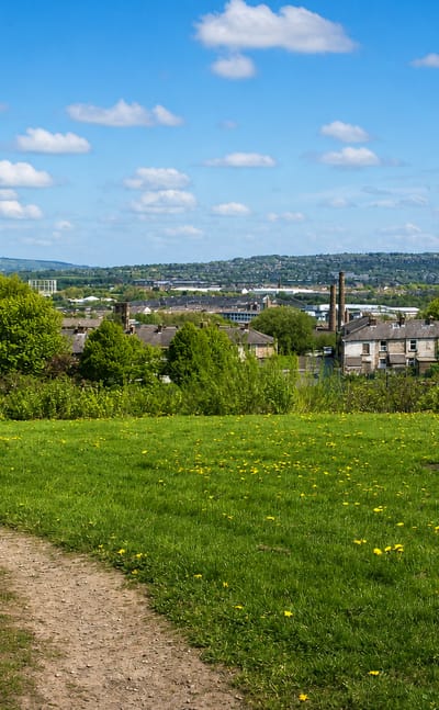 Grassy hilltop park path overlooking Burnley's rooftops and a mill chimney in the valley below, with distant hills