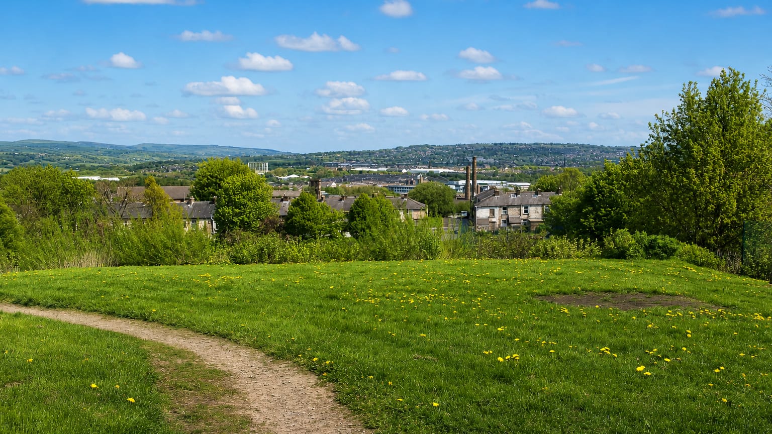 Grassy hilltop park path overlooking Burnley's rooftops and a mill chimney in the valley below, with distant hills