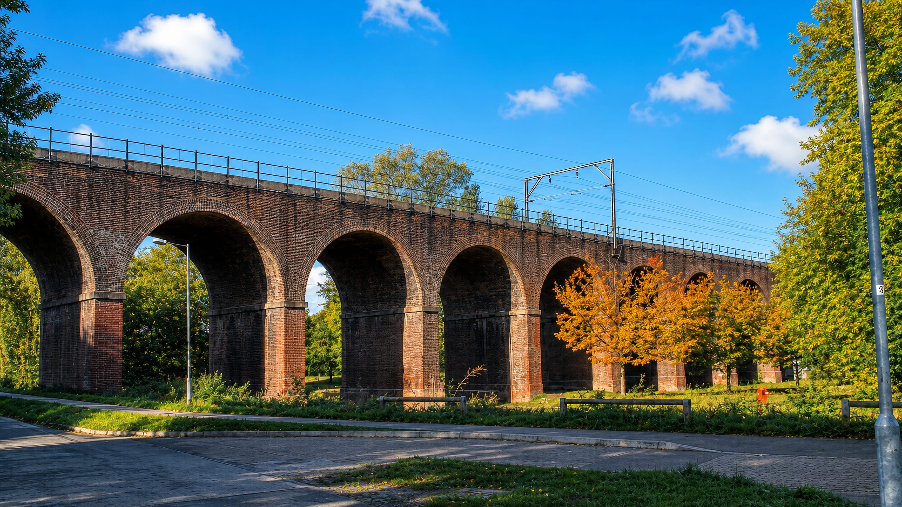 Red-brick railway viaduct arches carrying the main line above autumn trees and grass in Chelmsford, England.