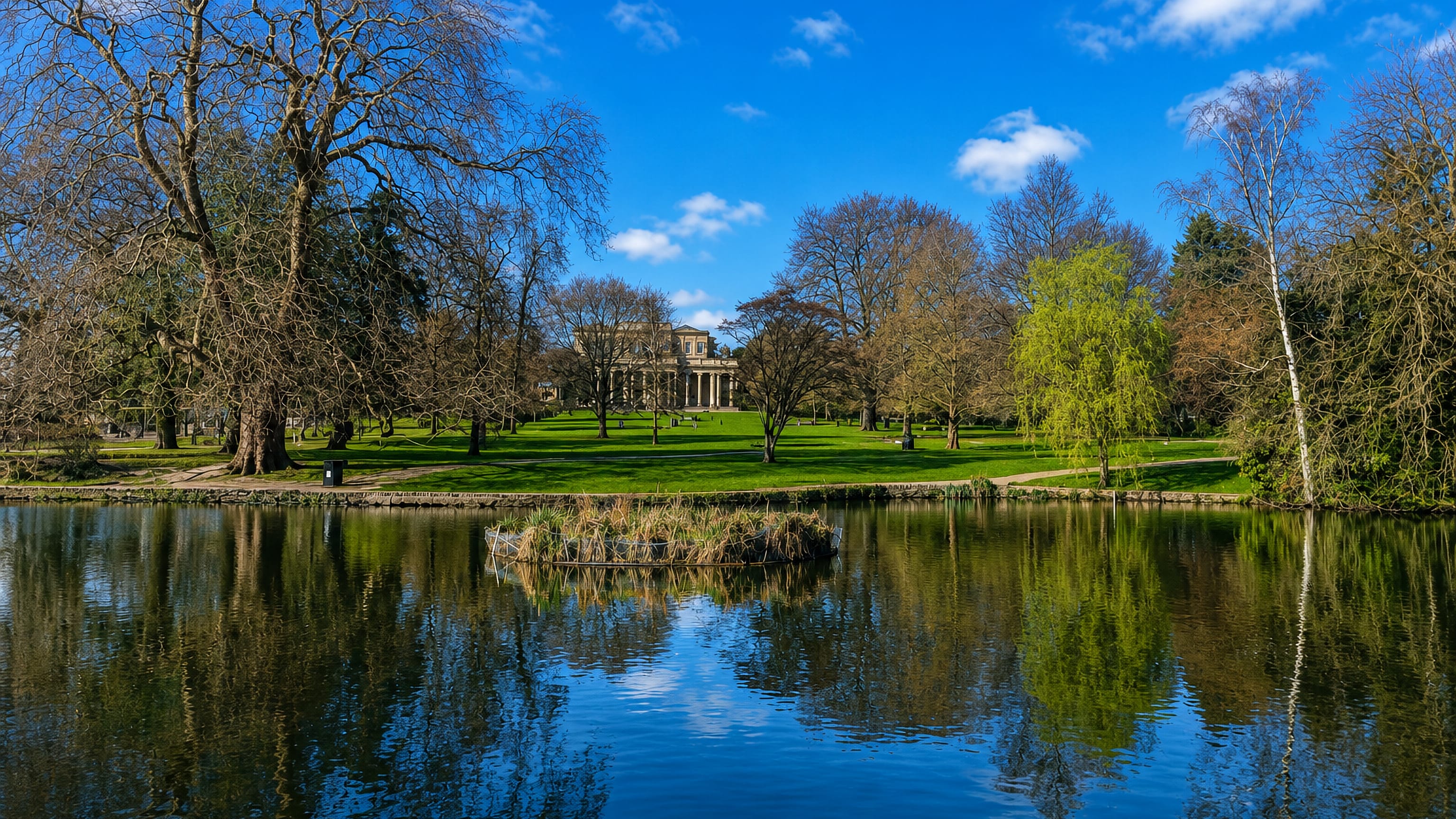 Pittville Park lake in Cheltenham reflecting bare trees, with the Regency Pump Room beyond the lawn.