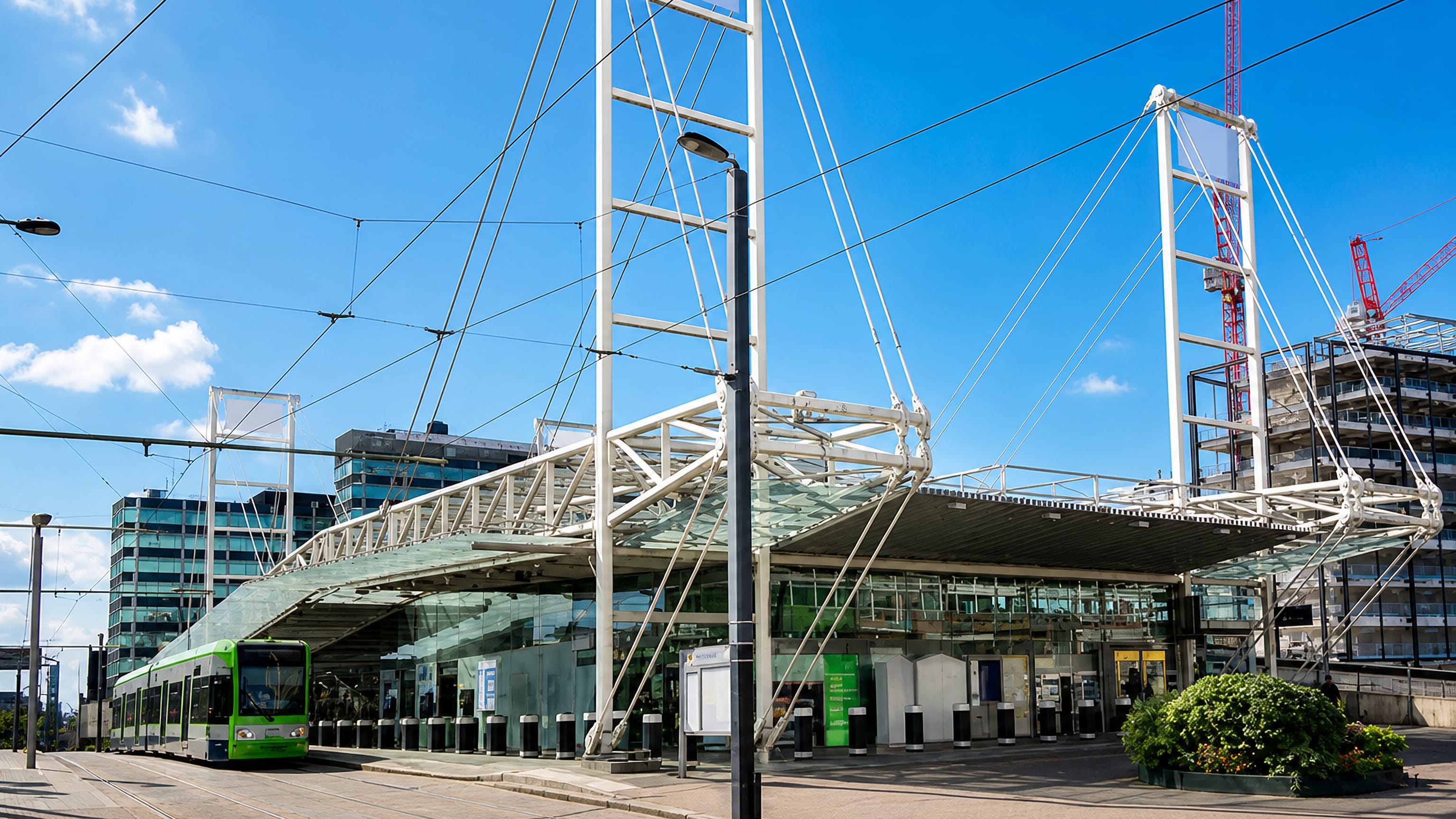 Green Croydon Tramlink tram beside the glass and steel canopy of East Croydon station under a blue sky.