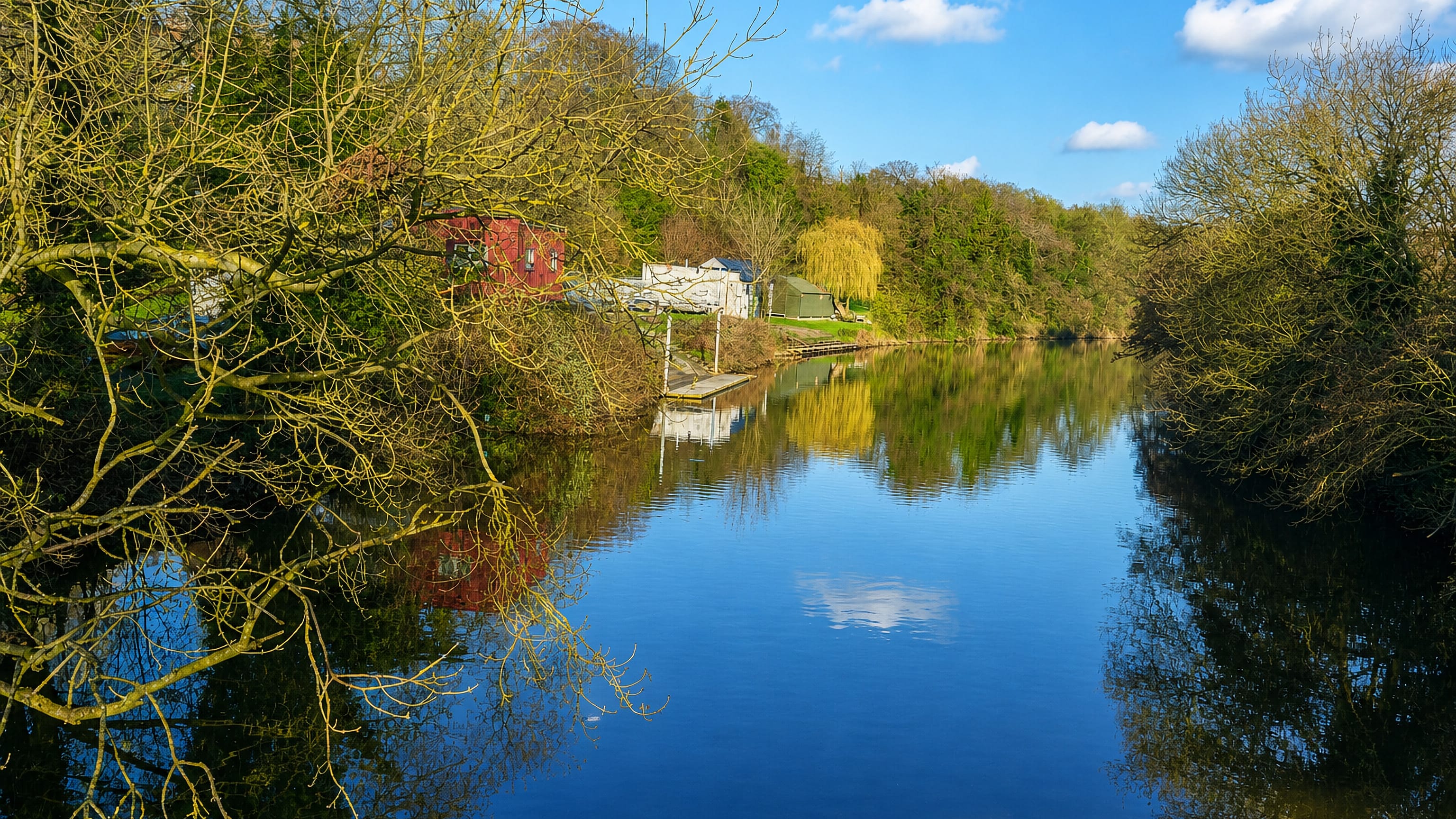 The River Derwent at Little Chester in Derby, wooded banks and small boathouses mirrored in still water
