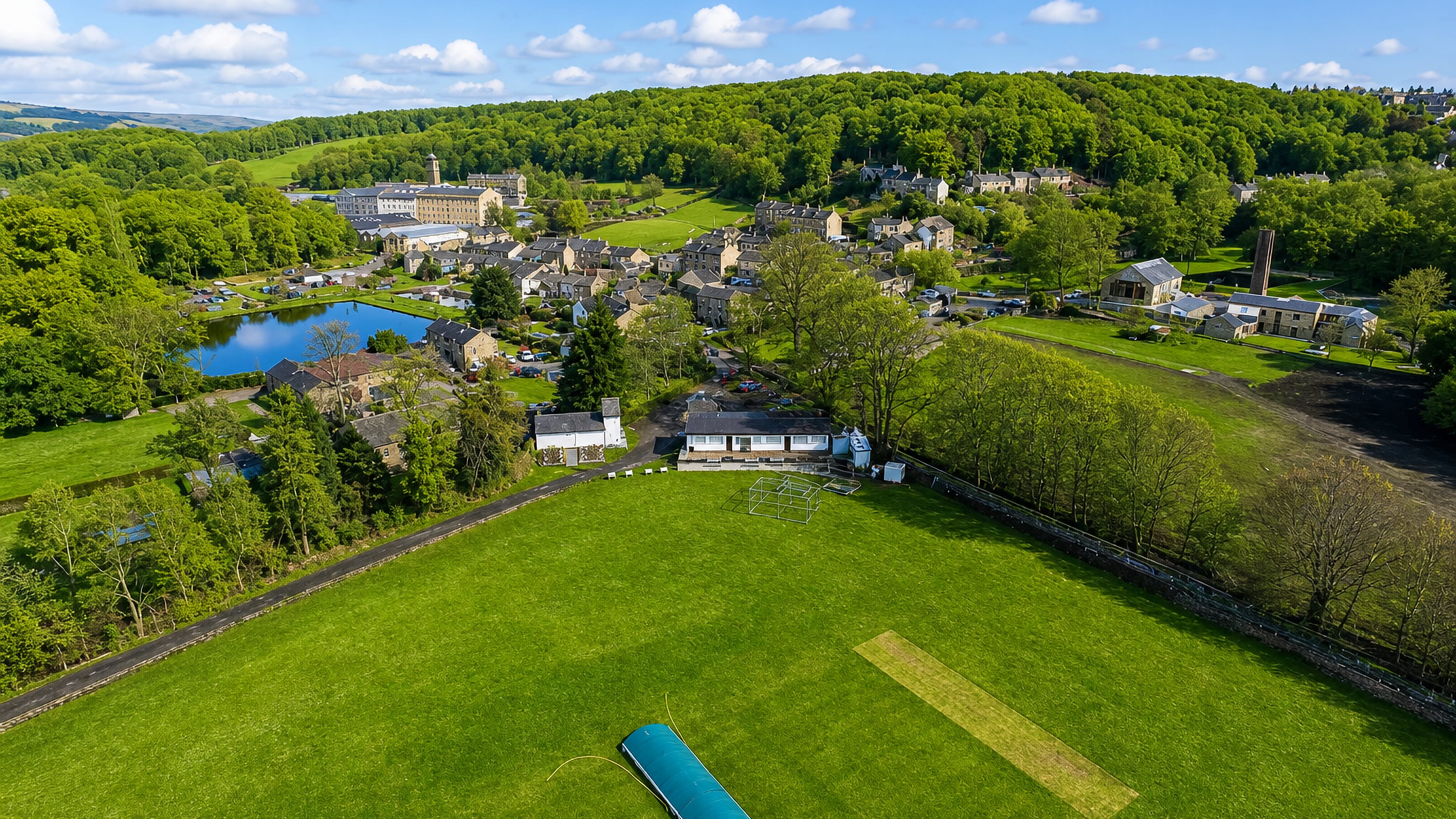 Aerial view of Armitage Bridge, Huddersfield, its cricket ground, stone village and mill below wooded hills.