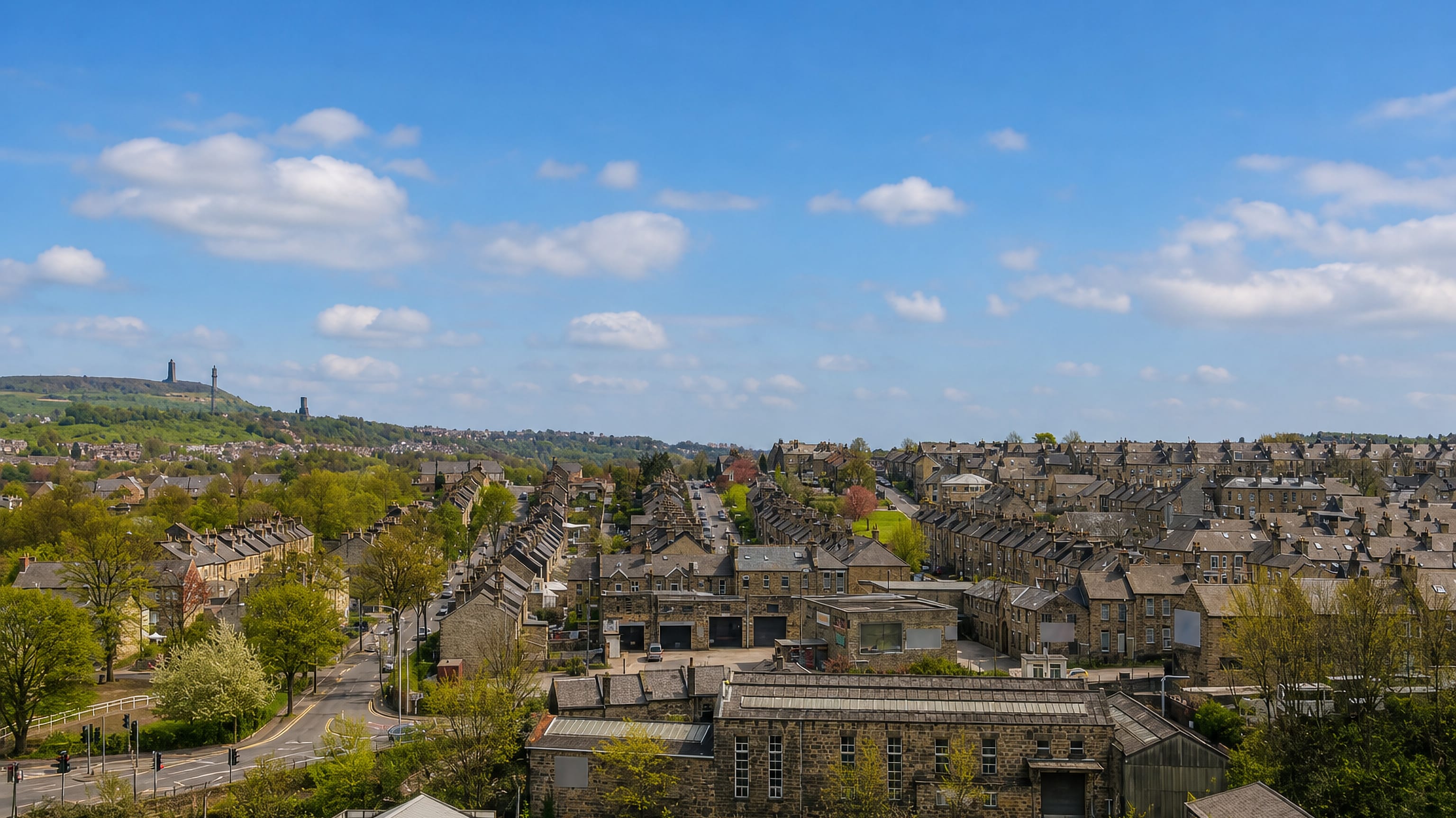 Stone terraced streets of Thornton Lodge, Huddersfield, rising below a wooded ridge topped by a distant hilltop tower.