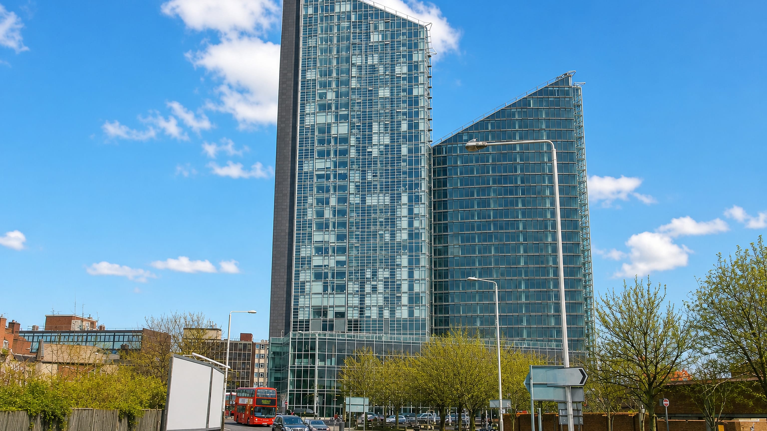 Twin angled glass towers rising above a main road with a red double-decker bus in Ilford, east London.