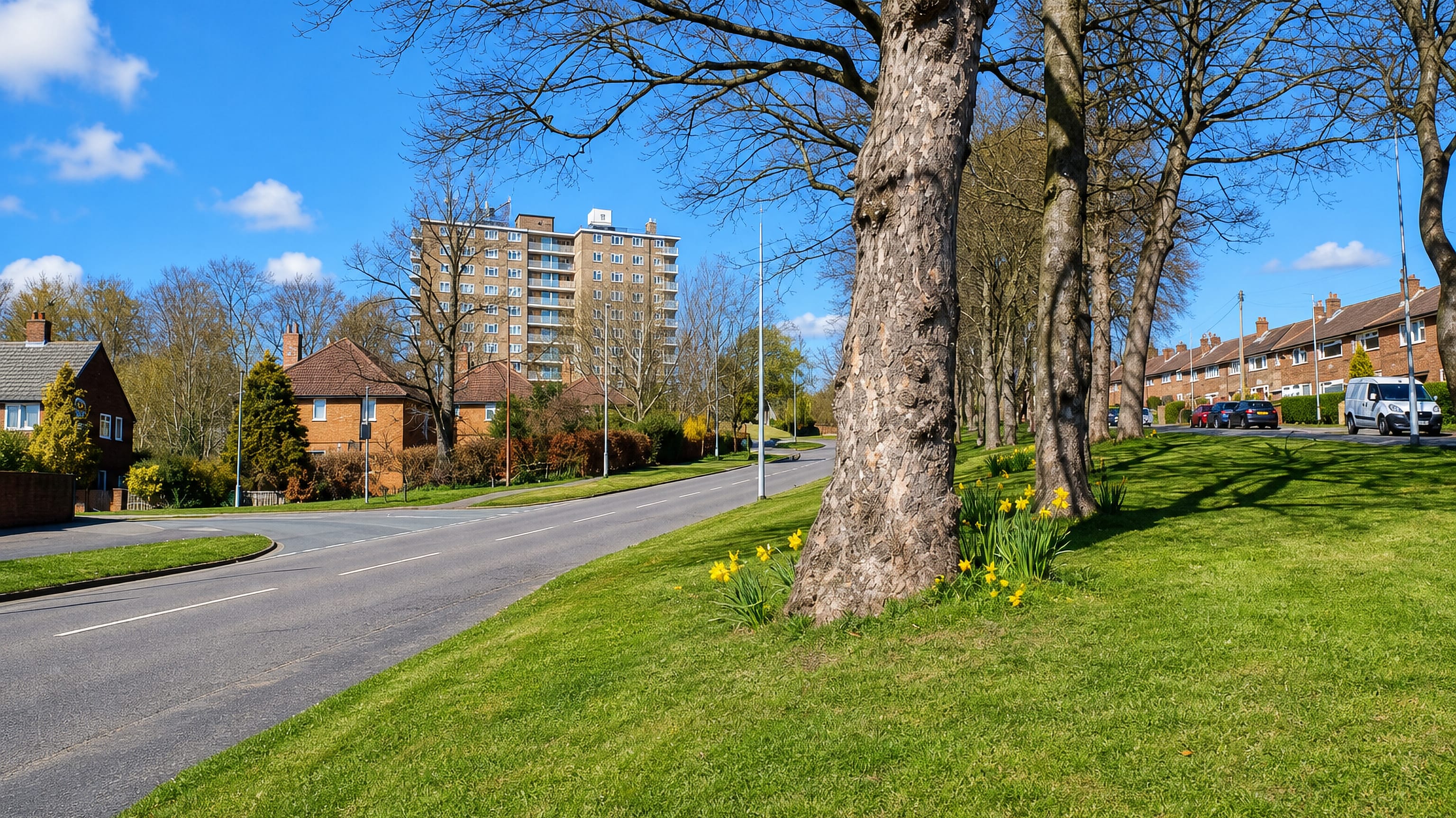 Grass verge and tree-lined road past brick houses and a tower block in Hawksworth, Leeds, in early spring.