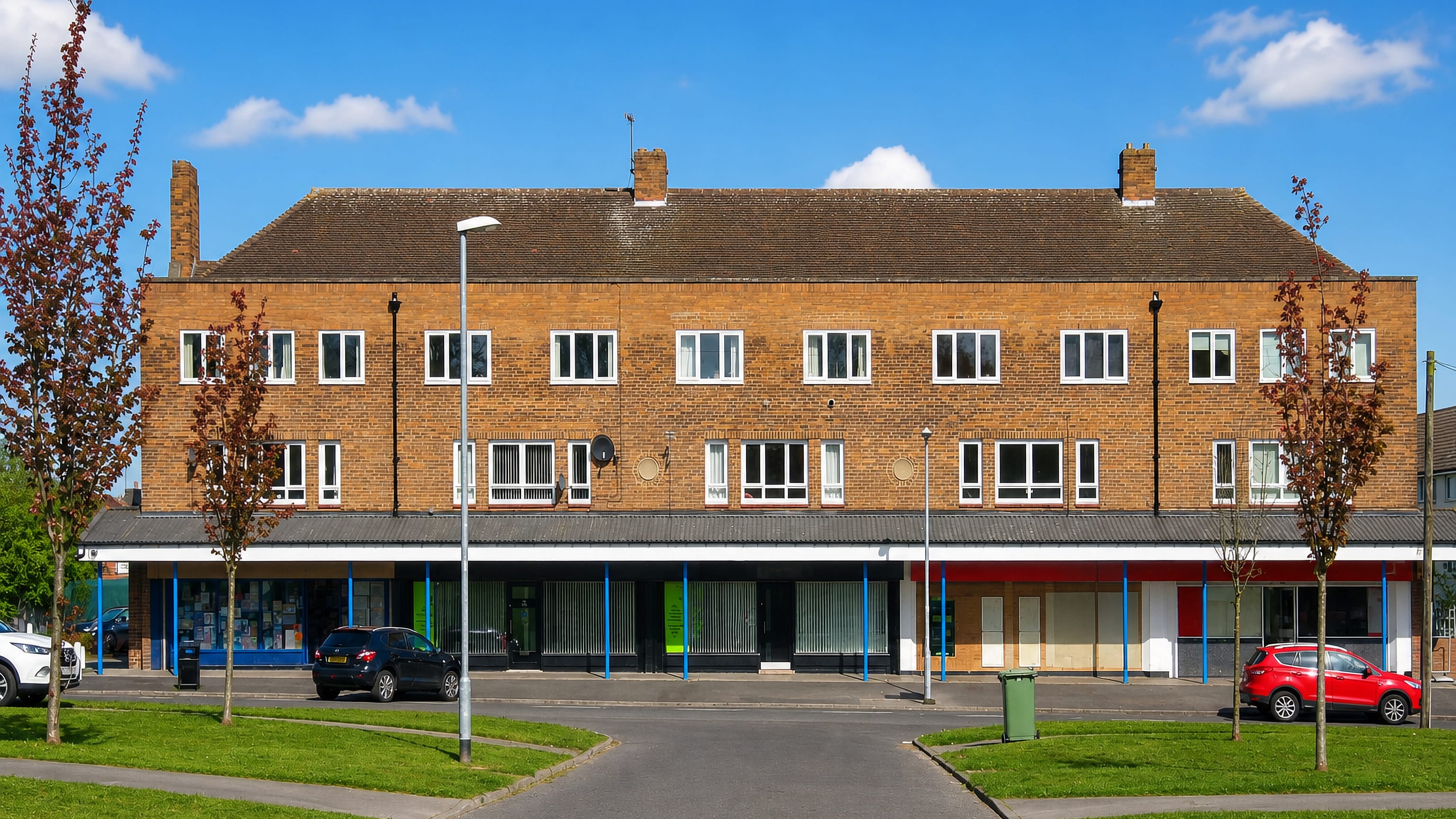 Sunlit brick shopping parade with flats above, fronting a green estate in Swarcliffe, east Leeds.