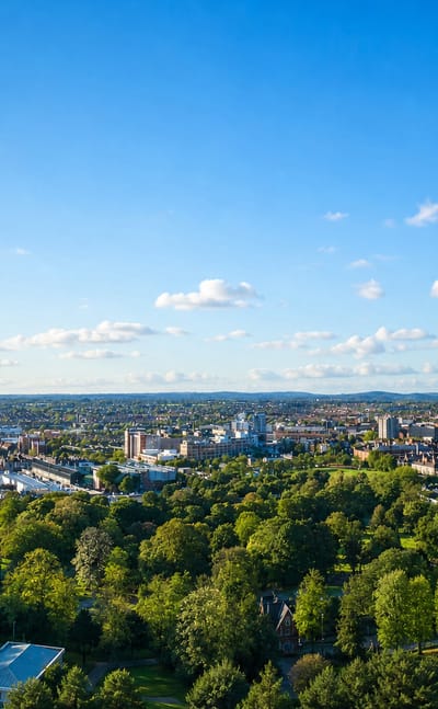 Aerial view over Leicester's leafy parkland toward a low city skyline under a wide blue sky.