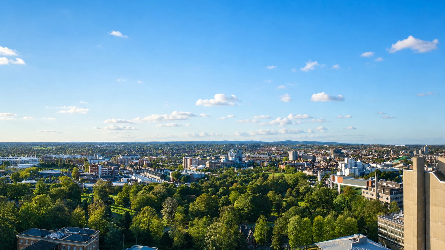 Aerial view over Leicester's leafy parkland toward a low city skyline under a wide blue sky.