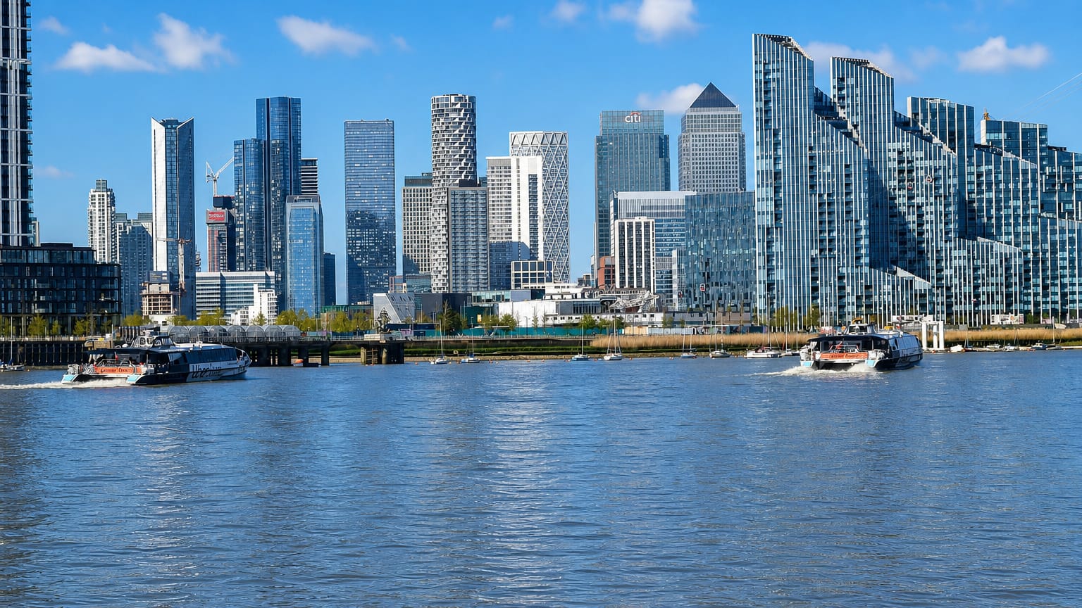 Canary Wharf's cluster of glass skyscrapers rising above the River Thames as boats pass, London.