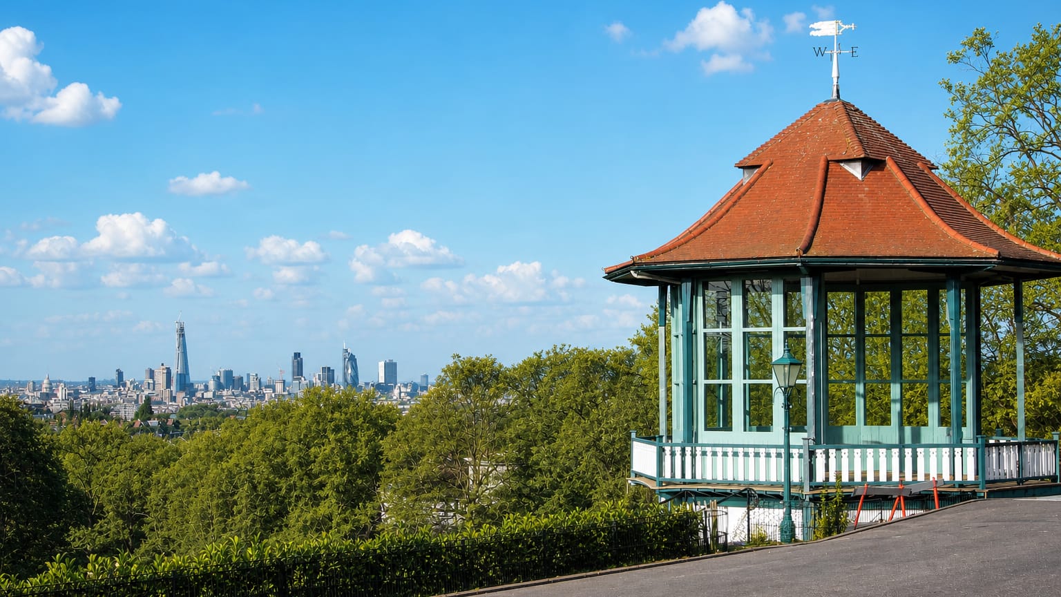 The ornate green bandstand in Horniman Gardens, Forest Hill, above treetops with London's skyline in the distance