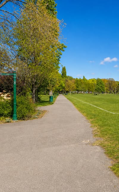 Sunlit tarmac path and green Victorian lamppost beside a wide park lawn under blue skies in Harold Wood, London.