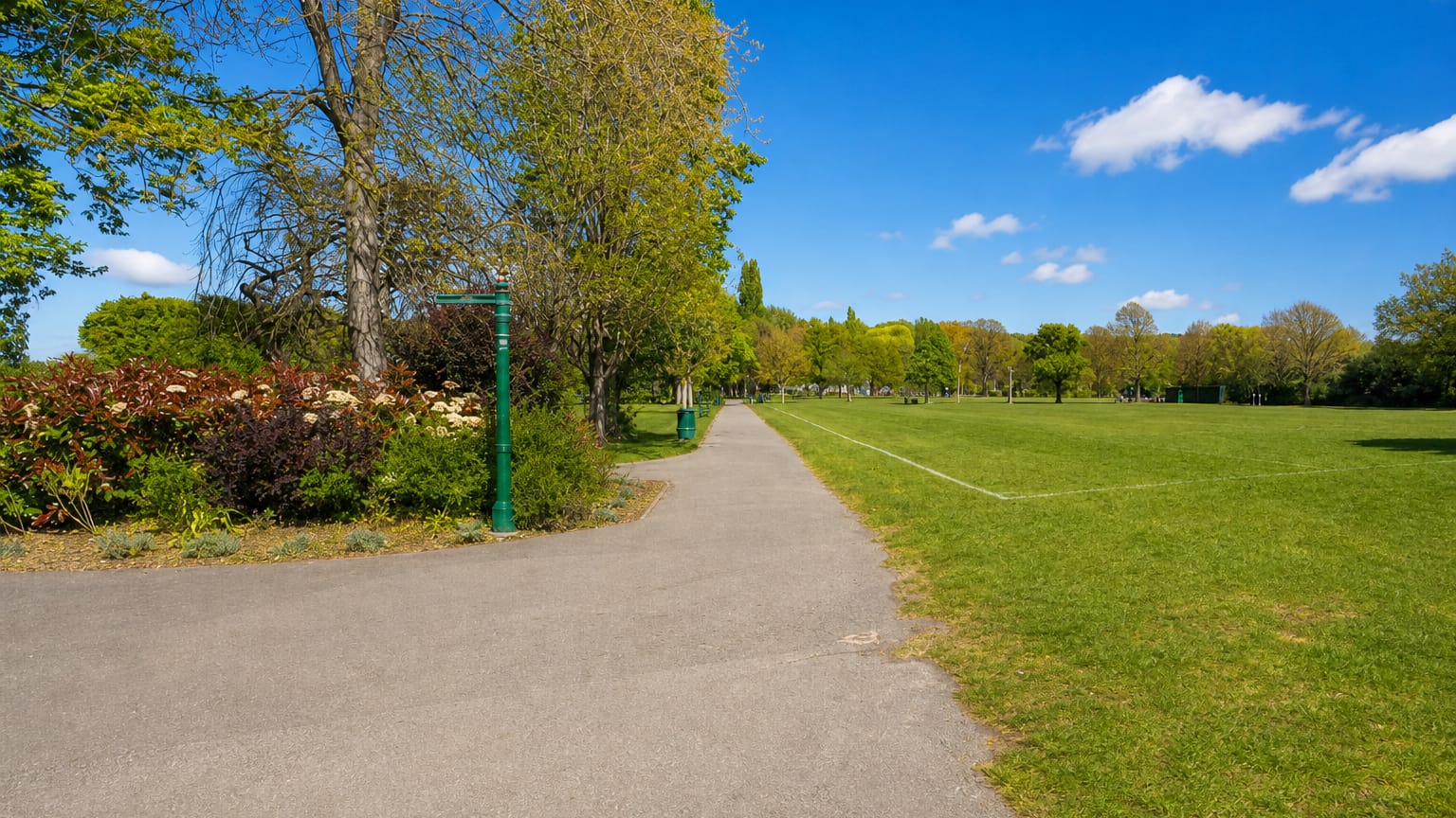 Sunlit tarmac path and green Victorian lamppost beside a wide park lawn under blue skies in Harold Wood, London.