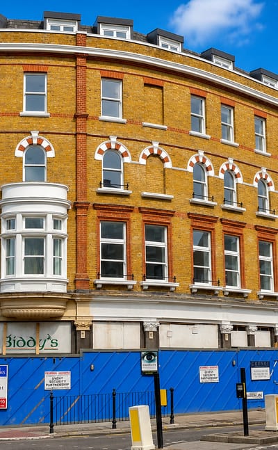 A yellow-brick Victorian corner building with shuttered shopfronts and to-let hoardings on a Kilburn street, London