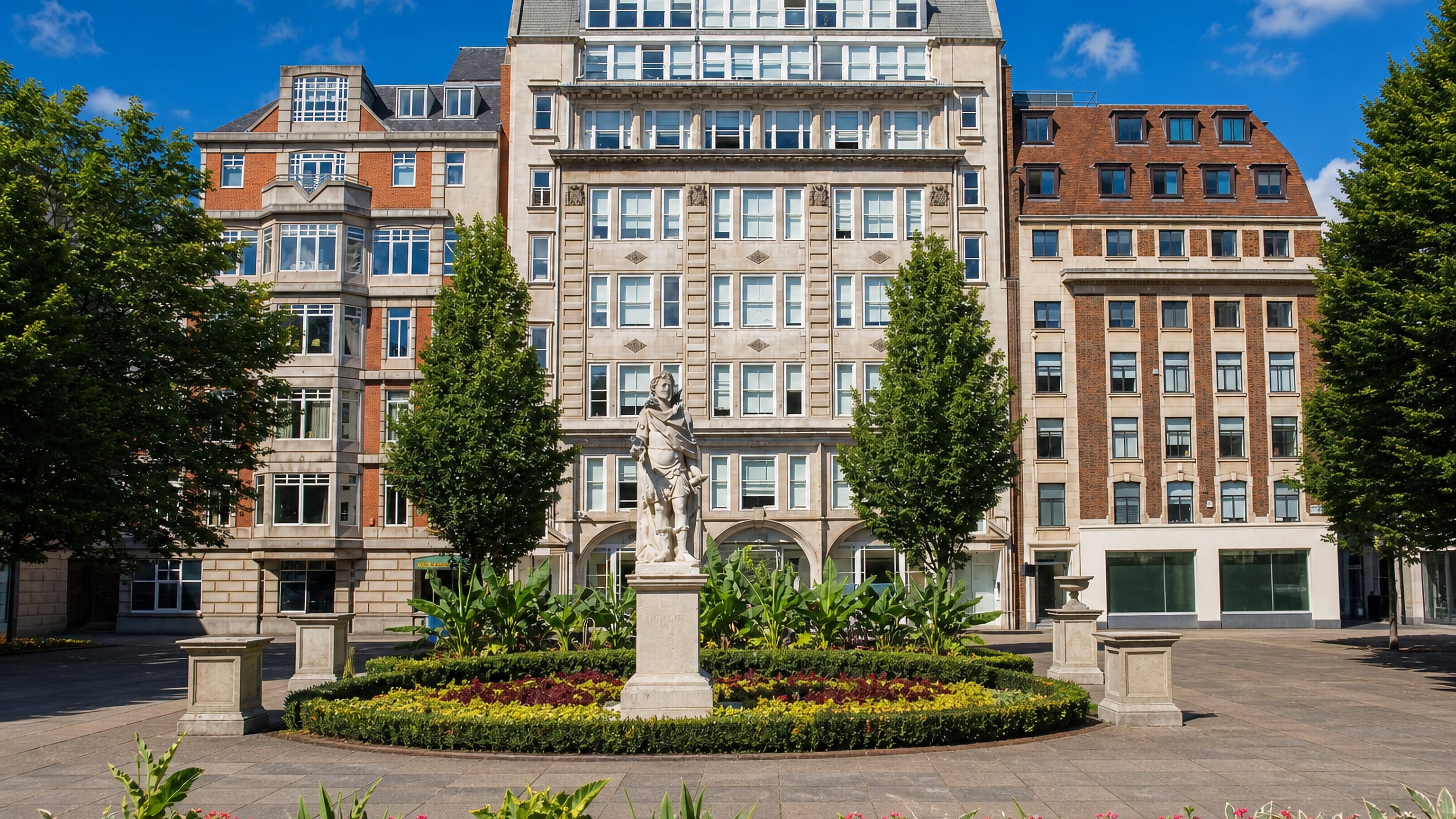 A statue and flowerbeds in a garden square in Soho, London, framed by brick and stone office blocks