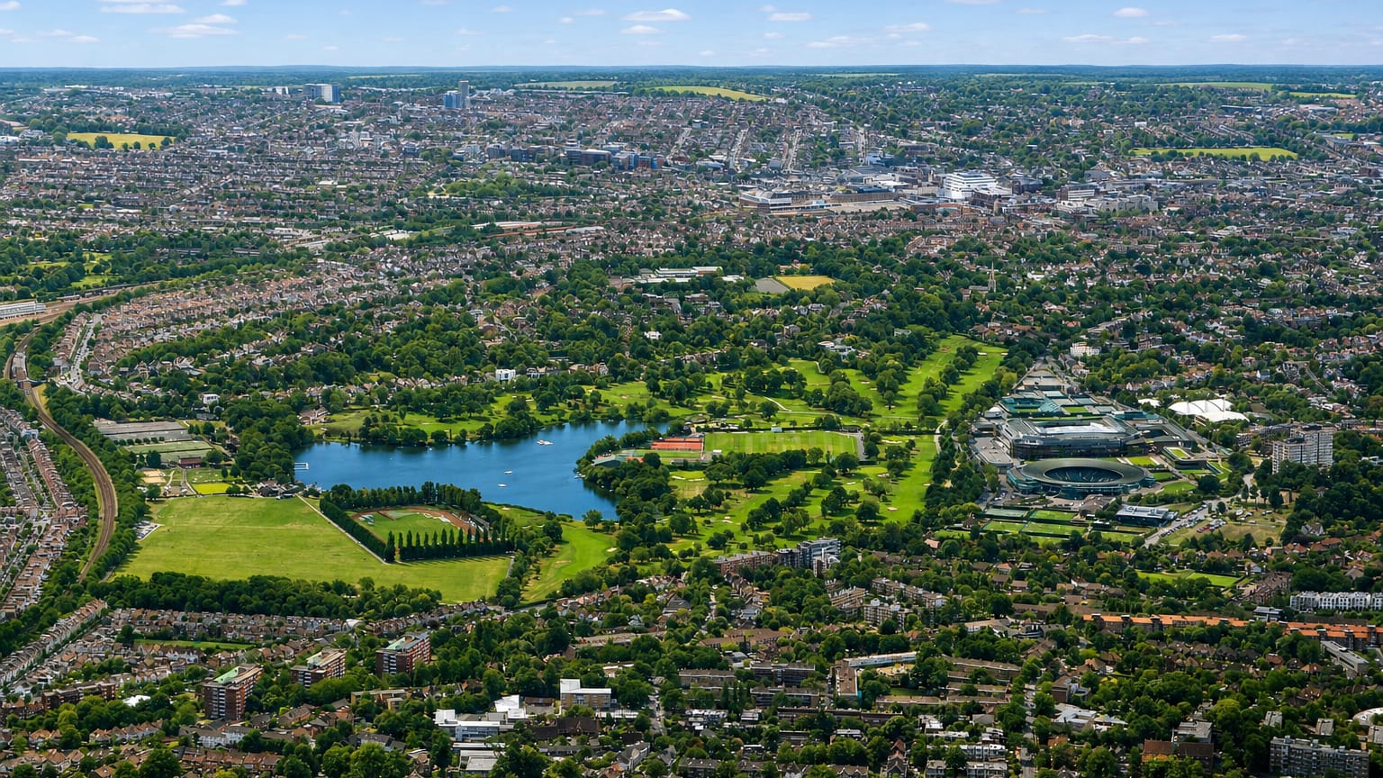 Aerial view over Wimbledon Park's lake and greenery toward the All England Lawn Tennis Club.