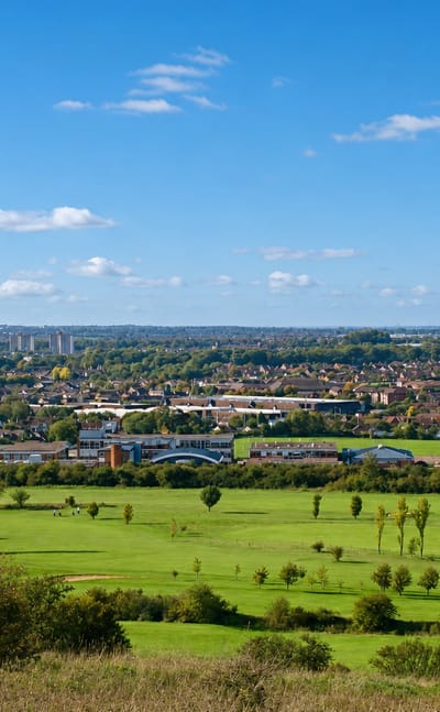Luton seen across green fields and a golf course in daylight, town rooftops and distant tower blocks on the horizon.
