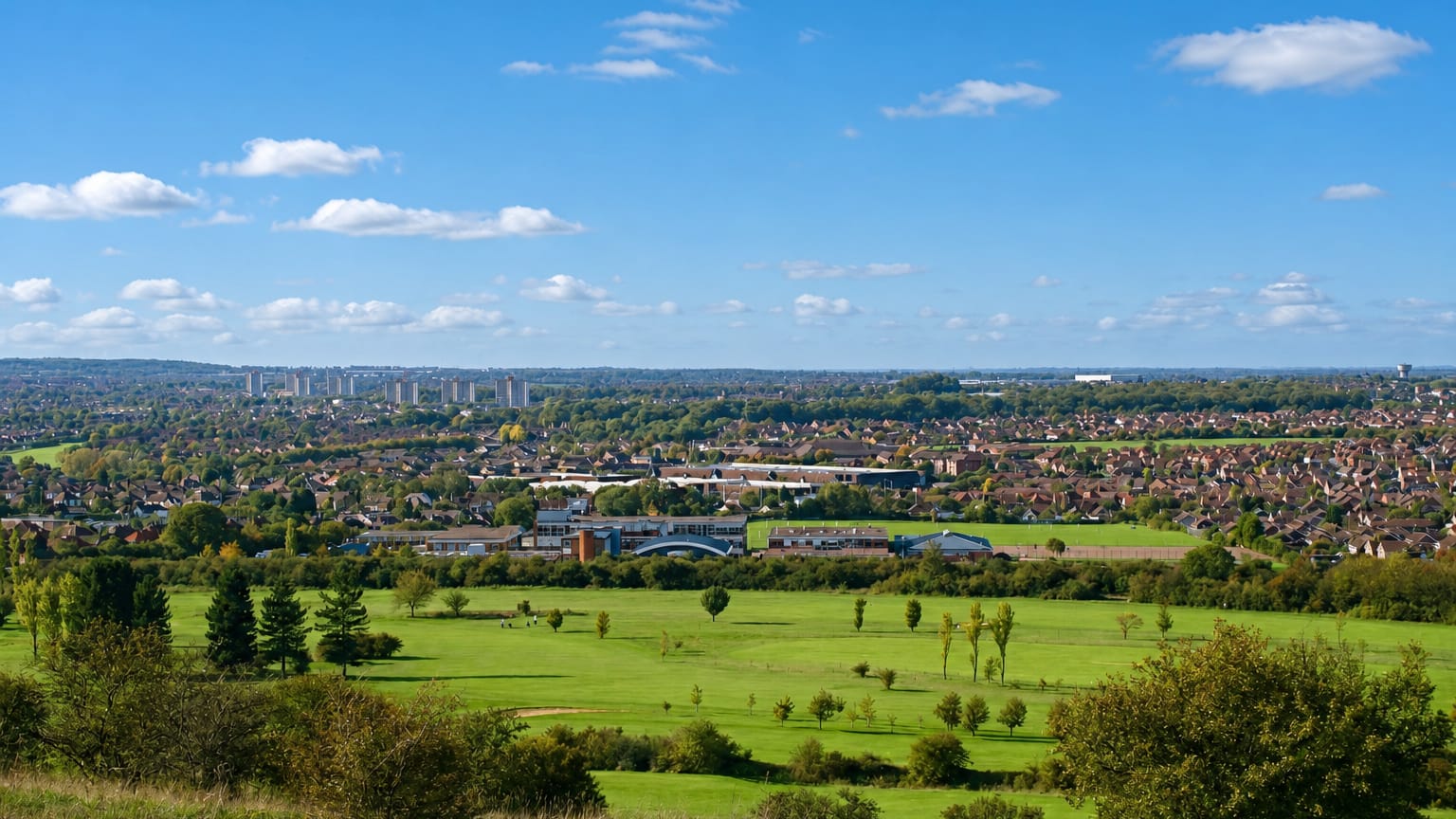 Luton seen across green fields and a golf course in daylight, town rooftops and distant tower blocks on the horizon.