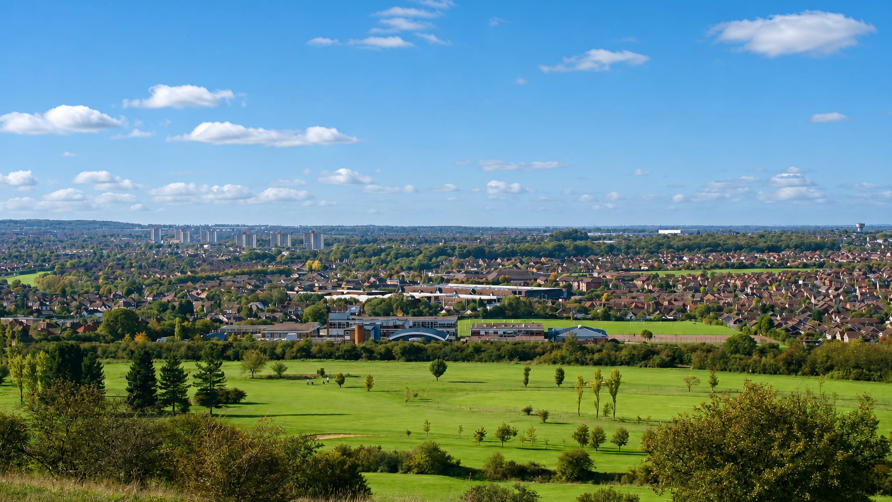 Luton seen across green fields and a golf course in daylight, town rooftops and distant tower blocks on the horizon.