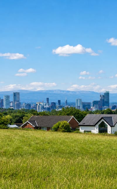 Manchester's cluster of skyscrapers on the horizon beyond a green meadow and suburban houses.