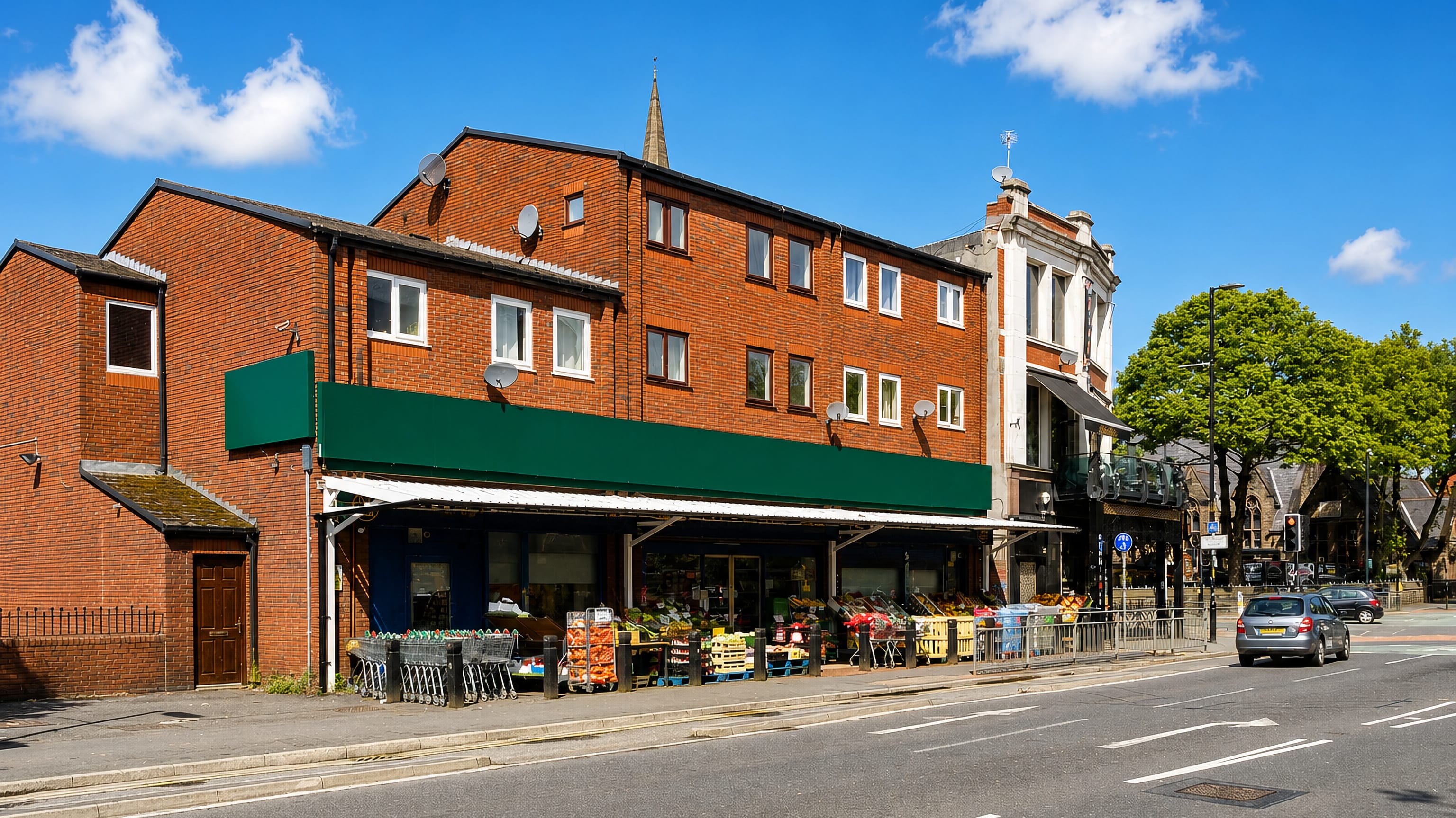 Red-brick shop parade with a greengrocer and church spire on the main road through Fallowfield, Manchester.