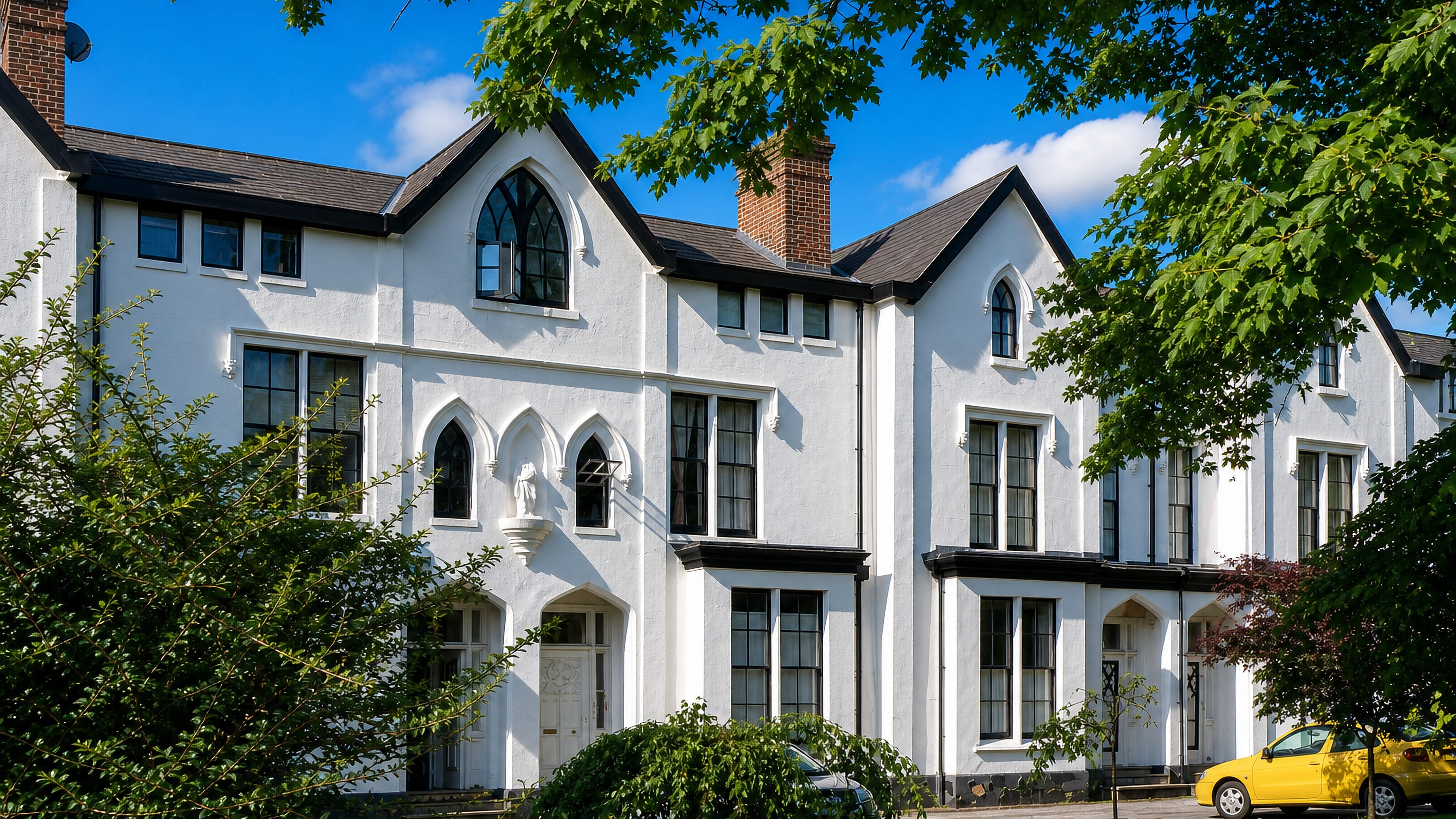 White Gothic Revival villas behind mature trees on a leafy street in Victoria Park, Manchester.