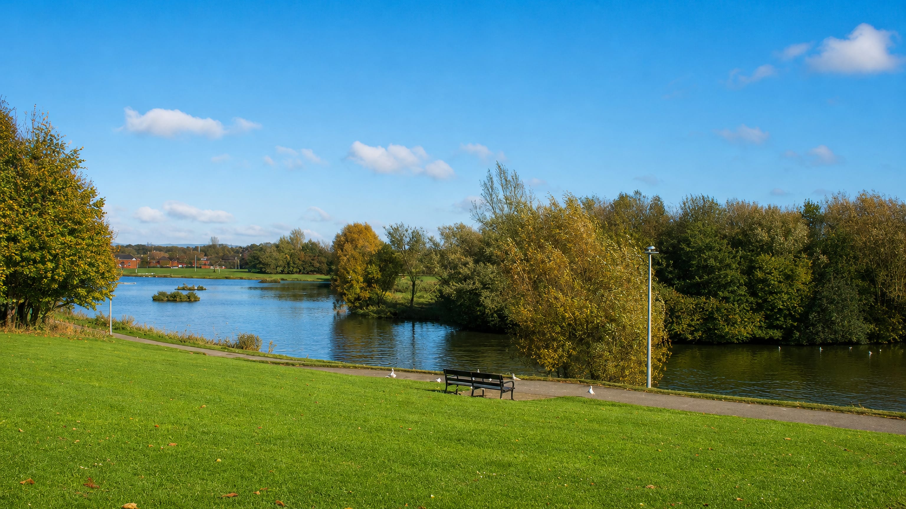 Hemlington Lake in Middlesbrough, with a bench, mown parkland and autumn trees along the water.