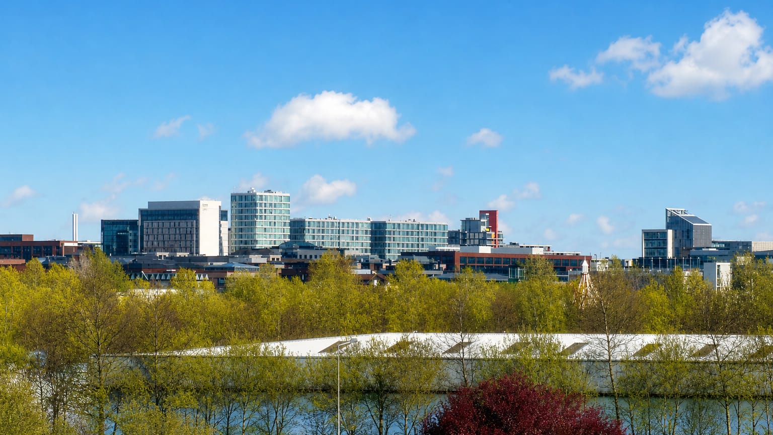 Modern glass office buildings of central Milton Keynes rise beyond parkland trees and a lake.