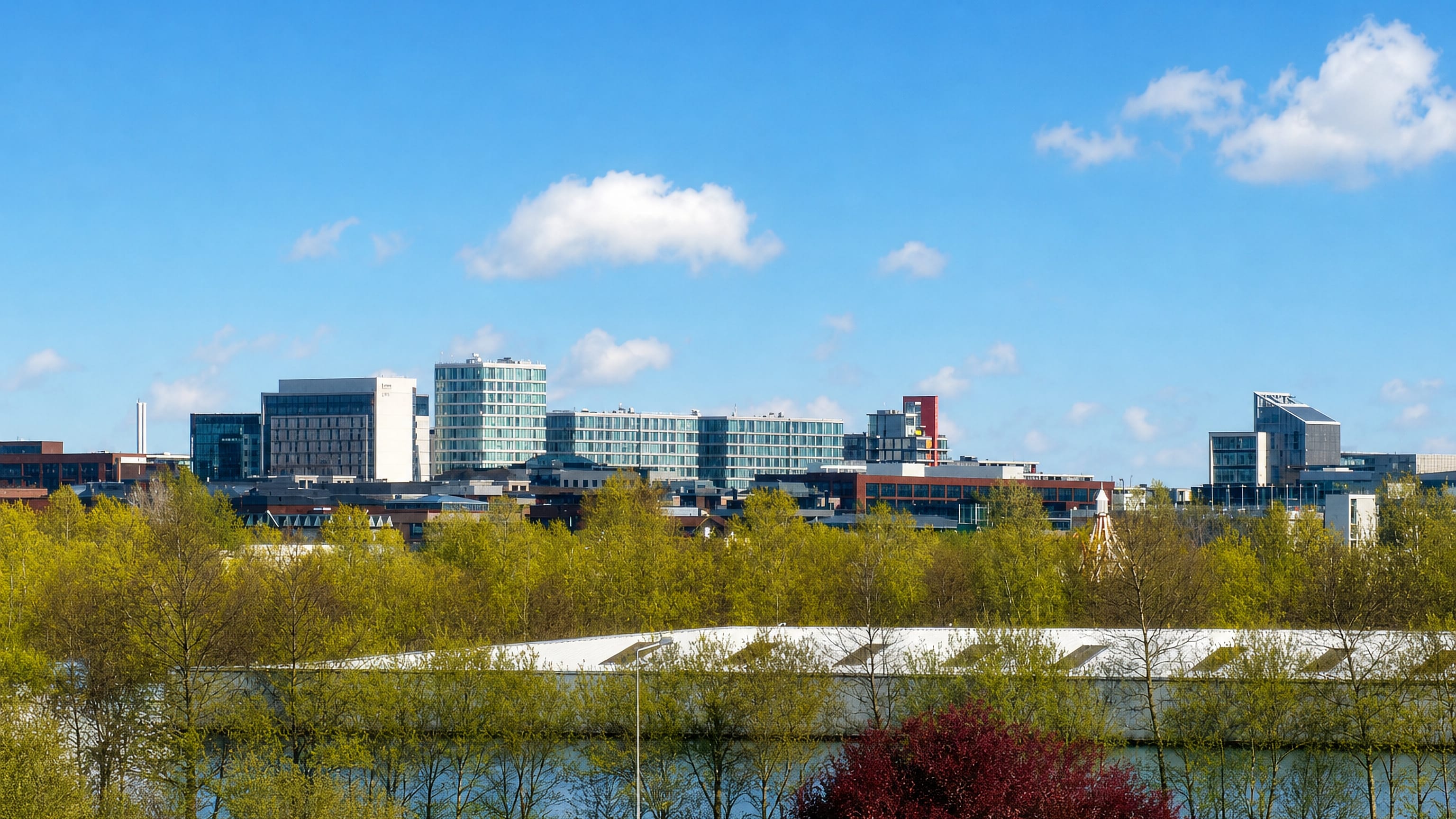 Modern glass office buildings of central Milton Keynes rise beyond parkland trees and a lake.