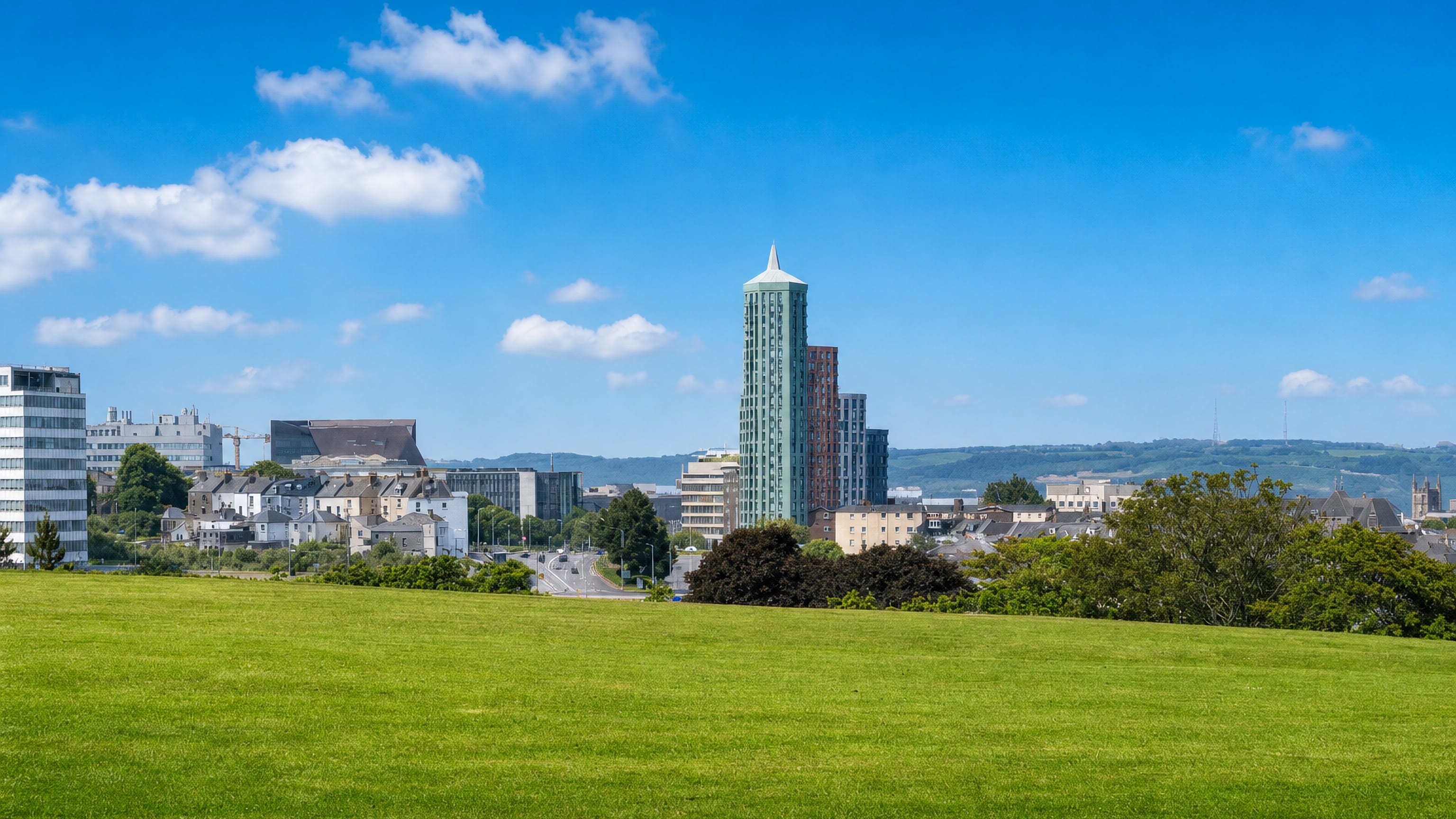 Plymouth city skyline seen across open parkland from Ford, with a tall spired tower above terraced streets.
