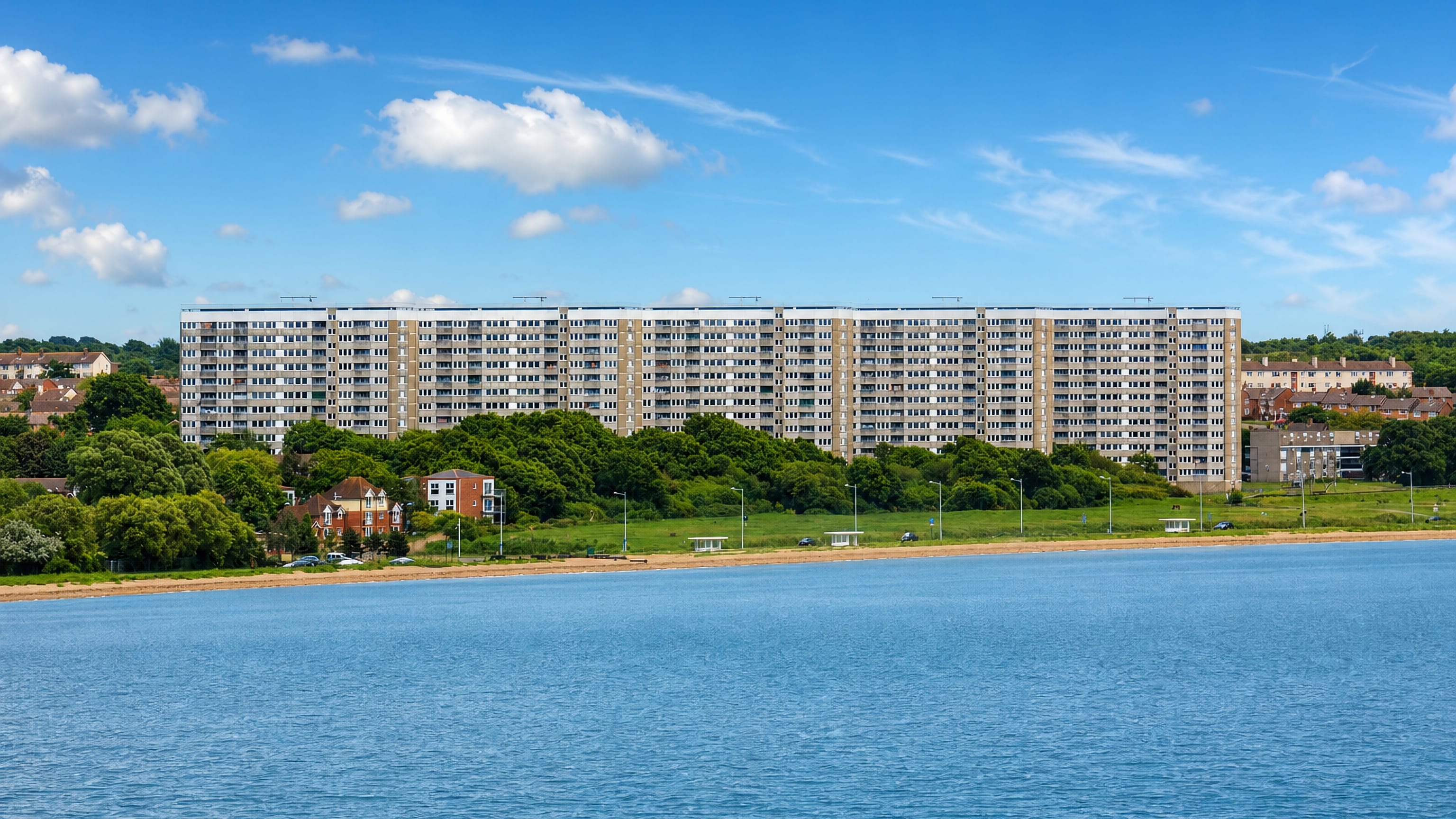 Long waterfront apartment blocks above the shingle shore at Woolston, Southampton, under a bright summer sky.