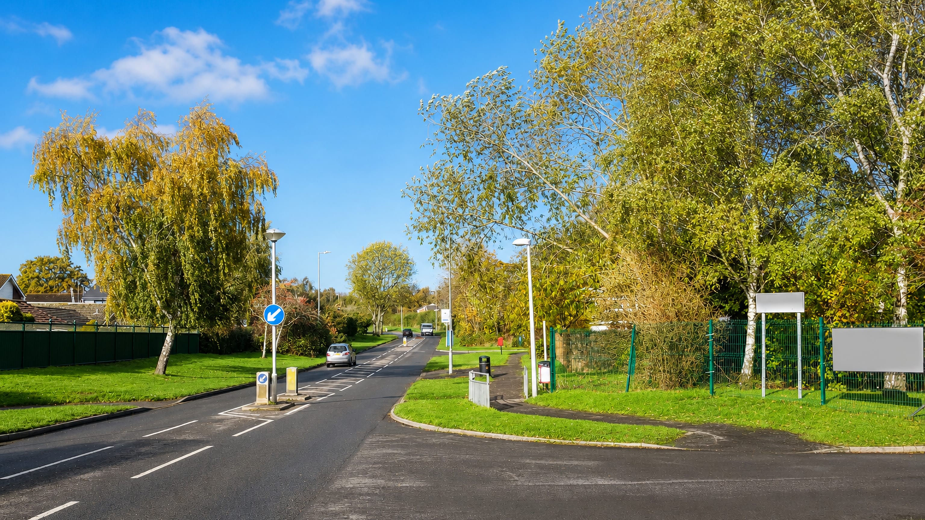 Tree-lined estate road with keep-left bollards and grass verges in Nythe, east Swindon, under a clear blue sky.