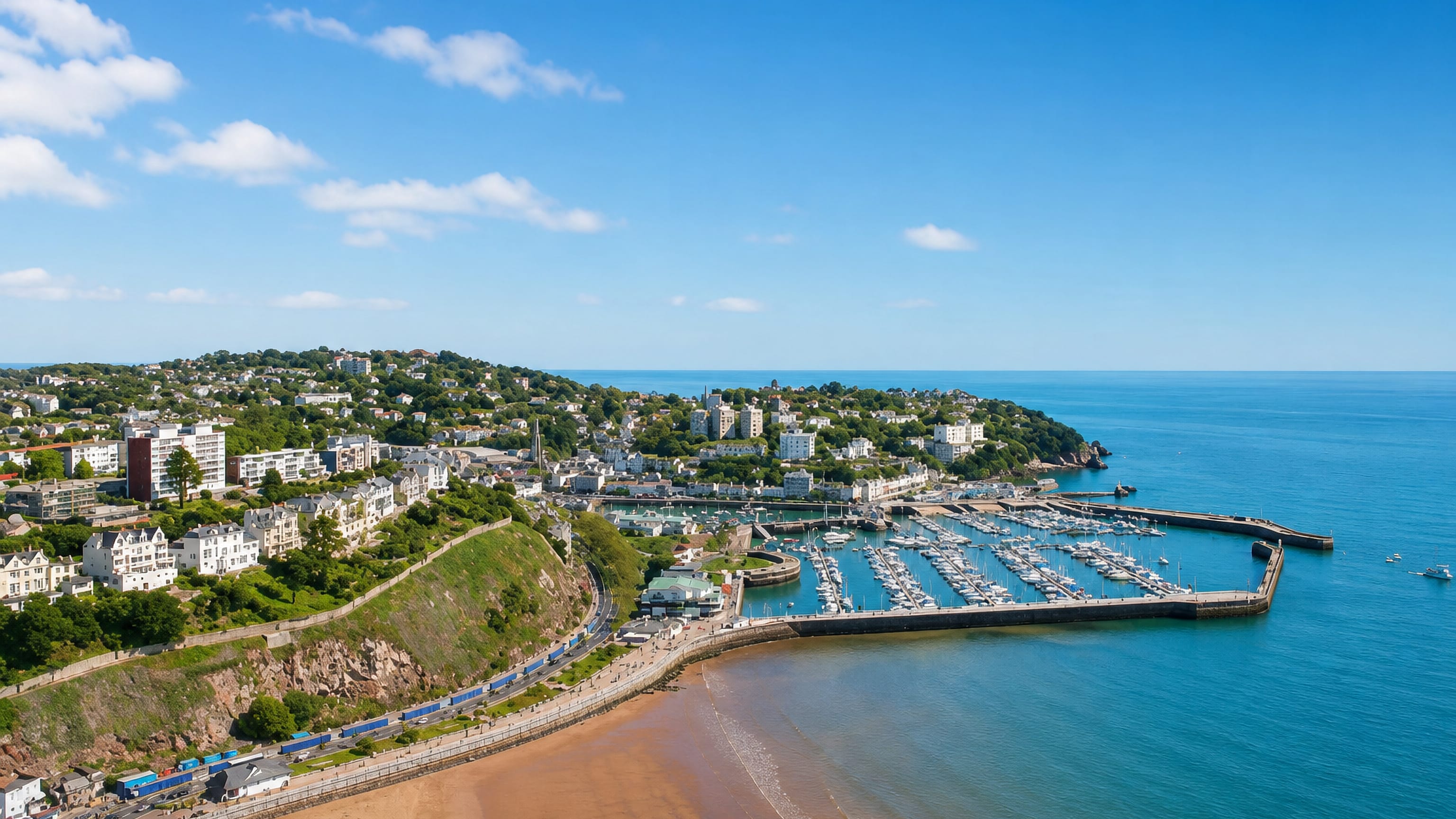 Torquay's harbour and marina below the wooded headland, with Torre Abbey Sands curving along the English Riviera coast.