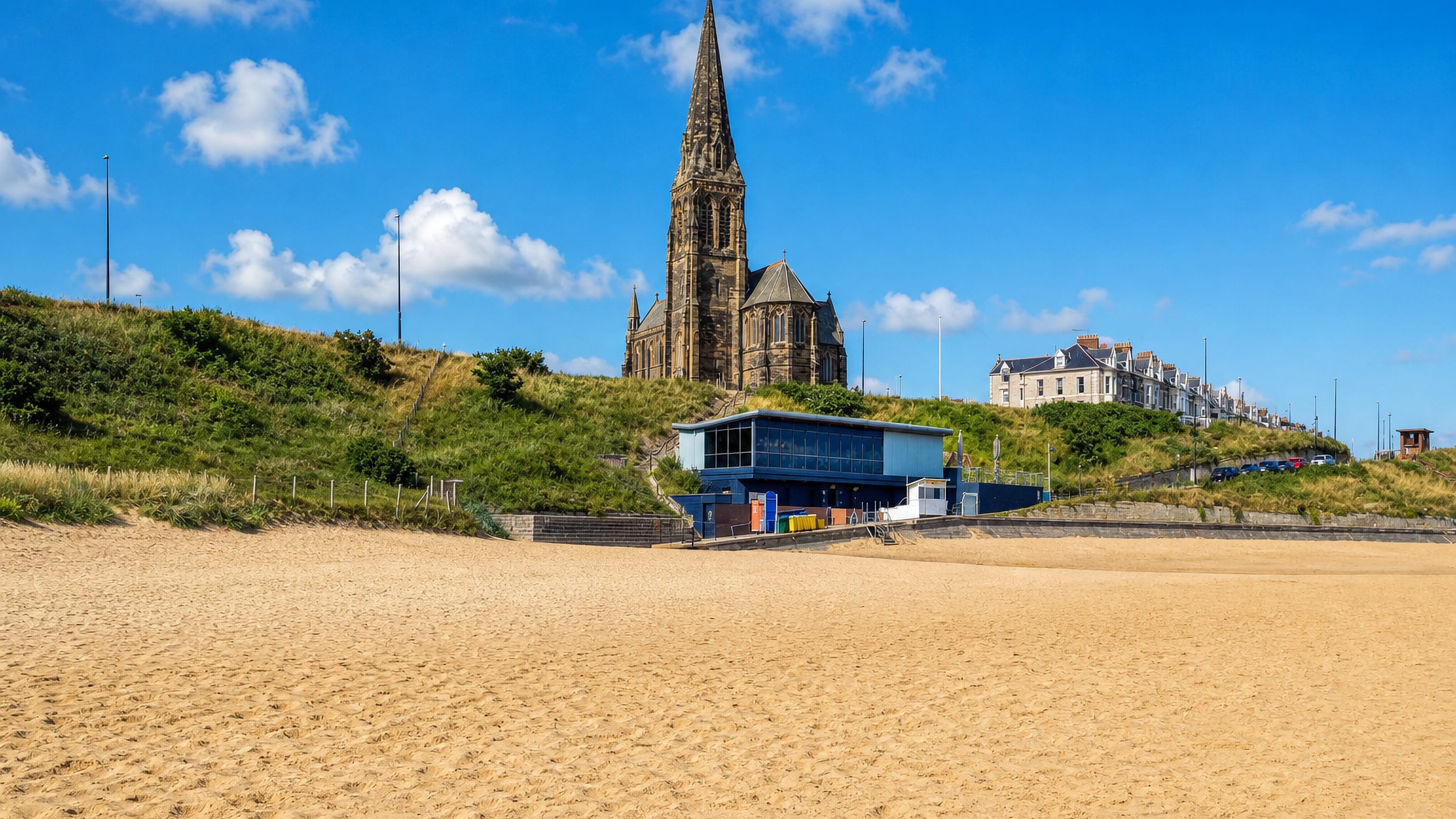 Sandy beach below a hilltop church spire and Victorian seafront terraces at Tynemouth, England.