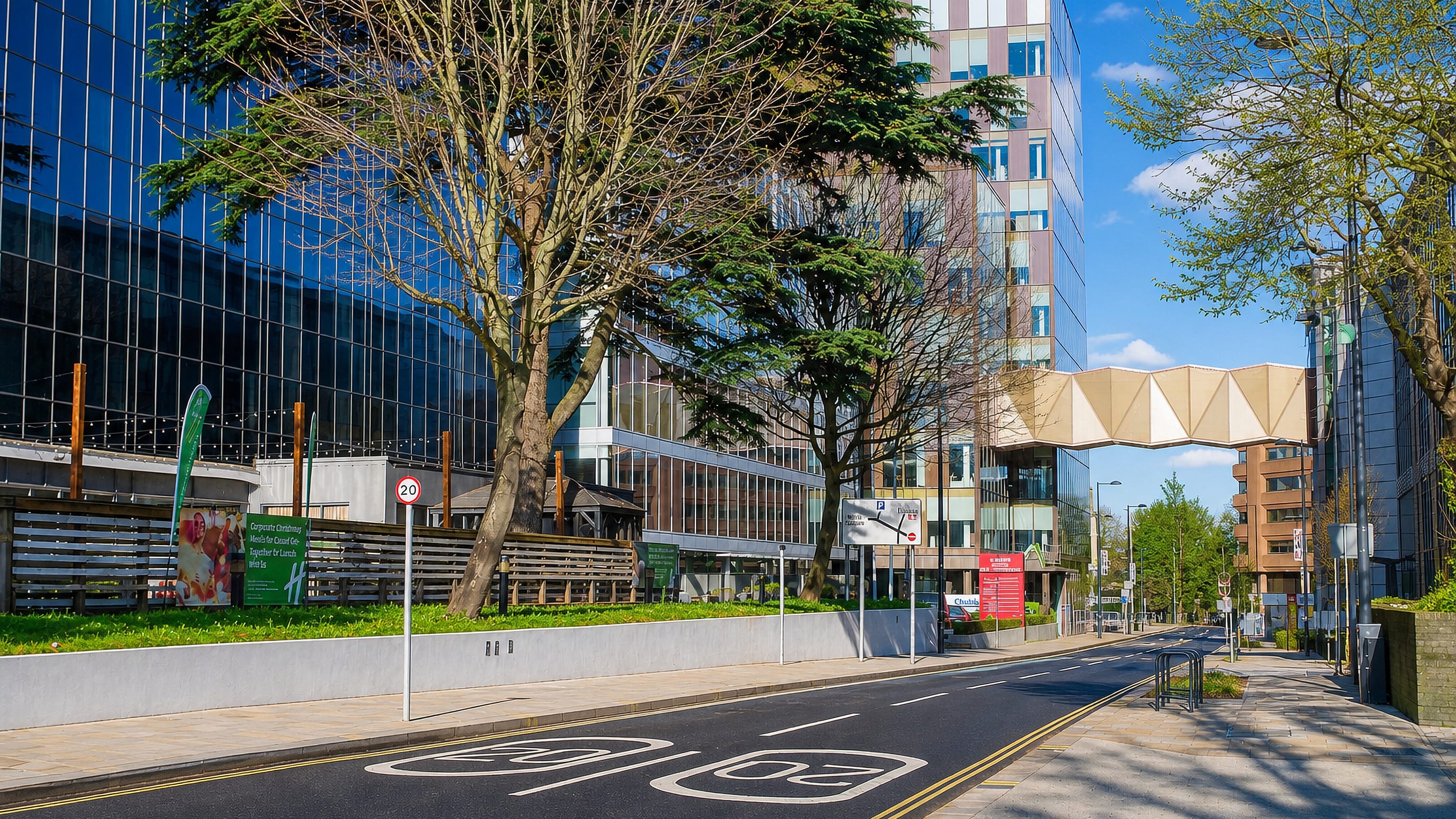 Glass office towers and a skybridge over a tree-lined business street in Watford, England.