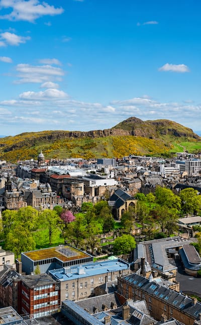 Edinburgh's old-town rooftops below the green slopes of Arthur's Seat and Salisbury Crags under blue sky.