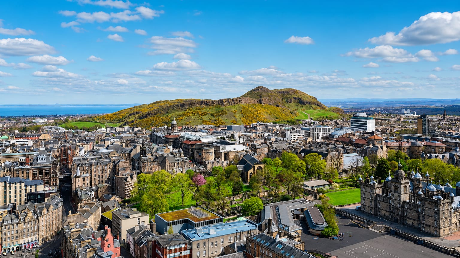 Edinburgh's old-town rooftops below the green slopes of Arthur's Seat and Salisbury Crags under blue sky.