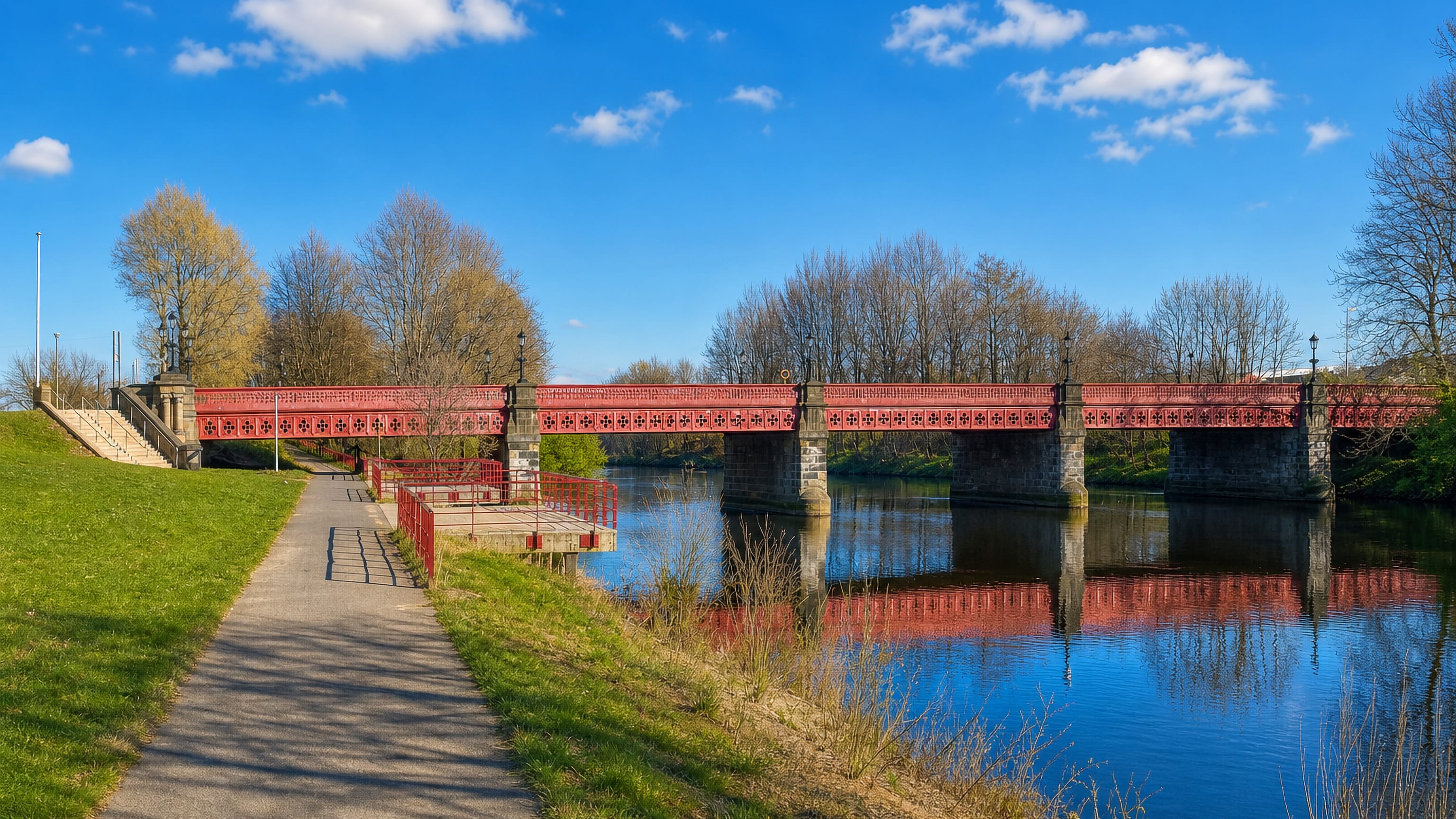 The red iron Dalmarnock Bridge crossing the River Clyde beside a riverside path in Glasgow.