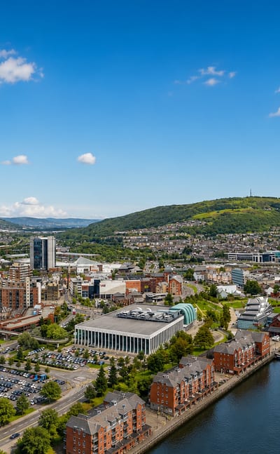 Aerial view of Swansea city centre and its marina with green hills rising behind under a blue sky.