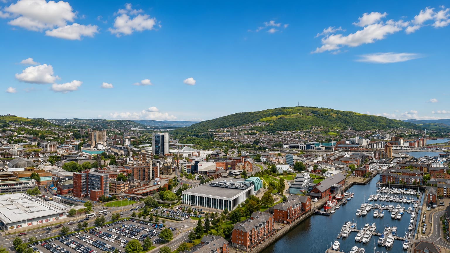 Aerial view of Swansea city centre and its marina with green hills rising behind under a blue sky.