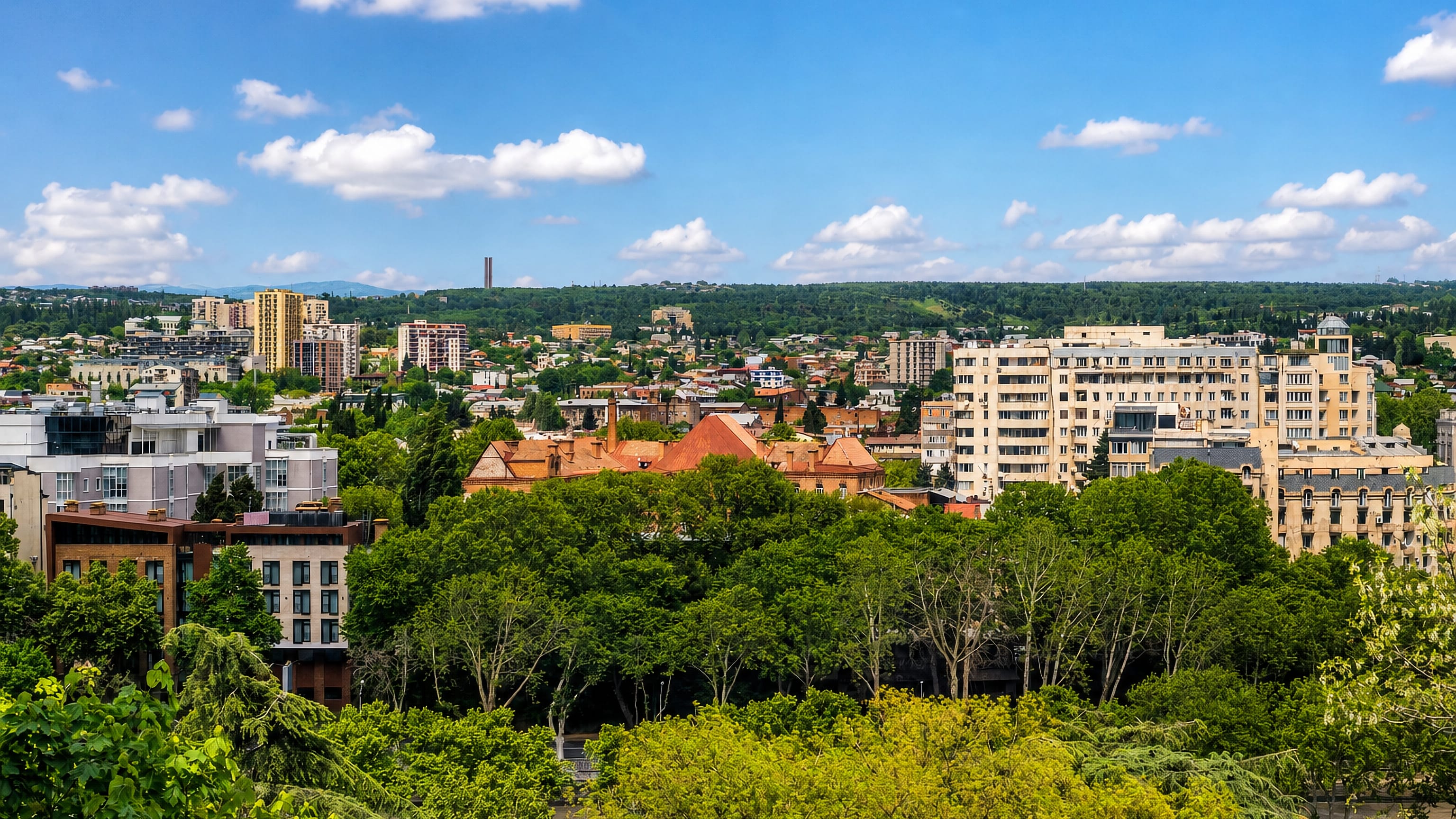 Tbilisi rooftops and apartment blocks above leafy treetops in Chughureti, with wooded hills beyond